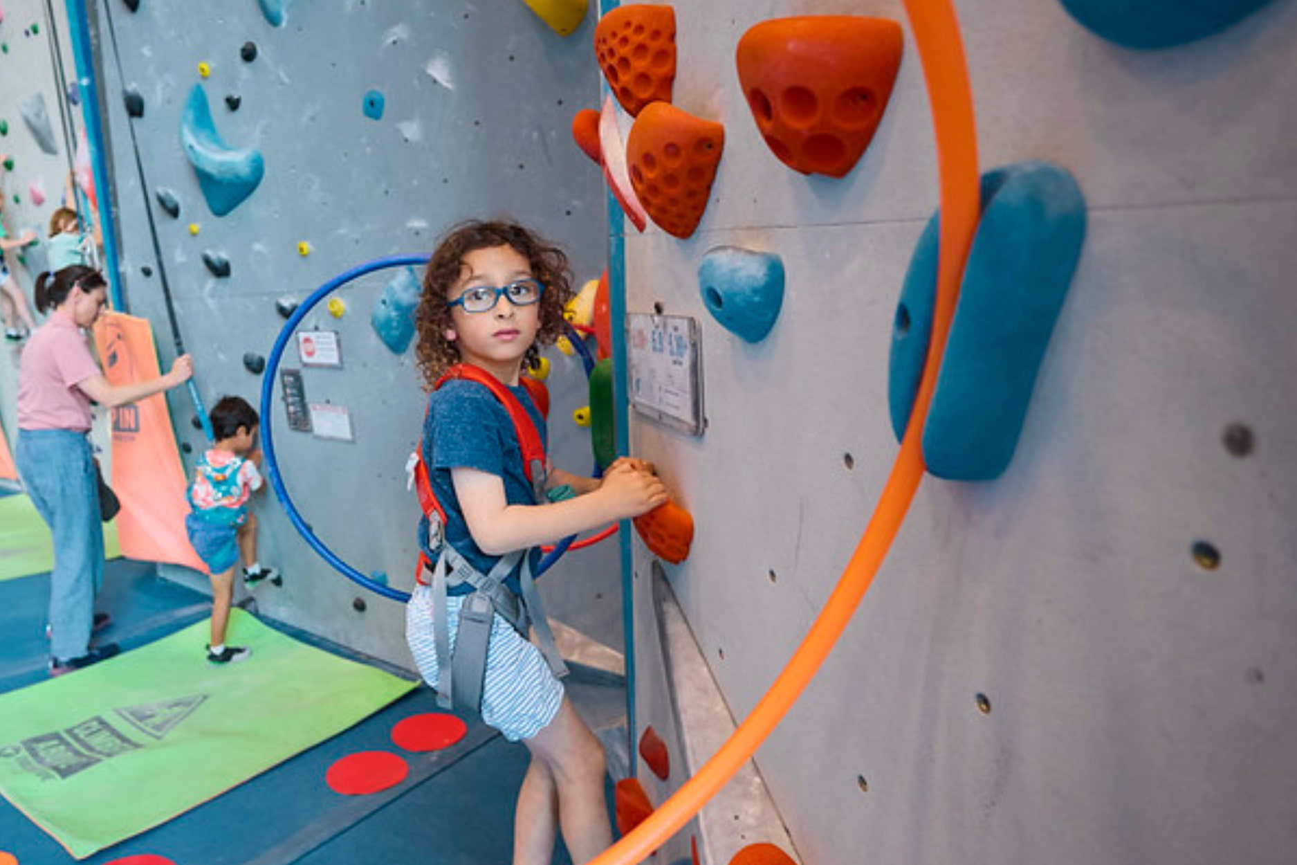 A young boy wearing glasses and a full body harness climbs through a hula hoop which is affixed to the climbing wall during a. youth program