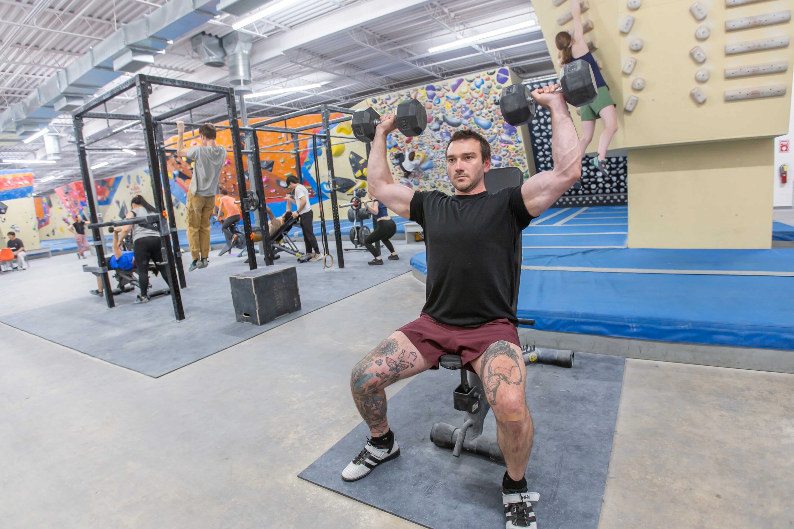 A seated man overhead presses with dumbbells as others train and work out in the backgroun.