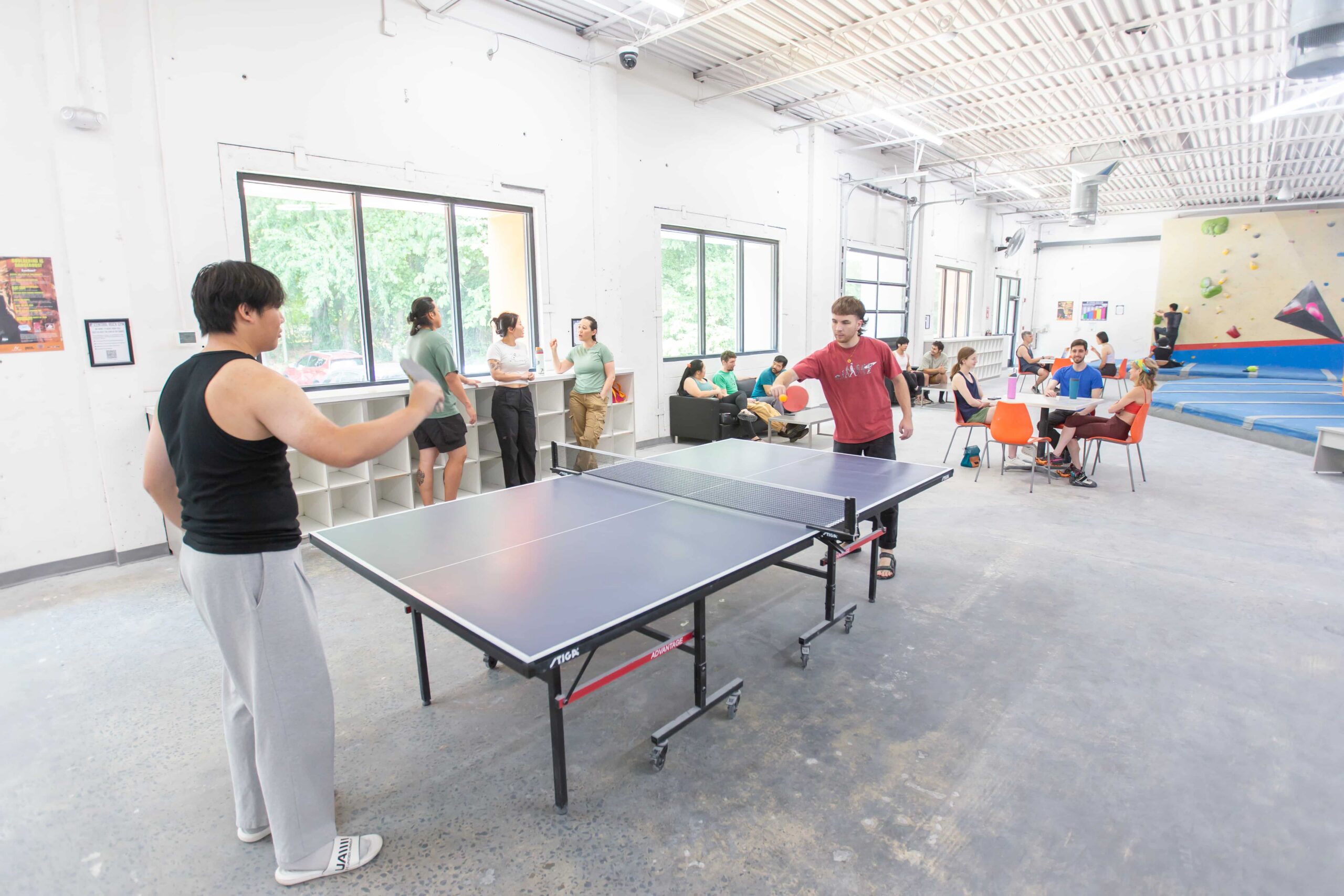 Two men play ping pong together. In the background people are socializing by the cubbies and sitting at tables together.