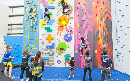 Children climb colorful, indoor climbing walls with geometric shapes and patterns. Staff members supervise, creating a lively and playful atmosphere.