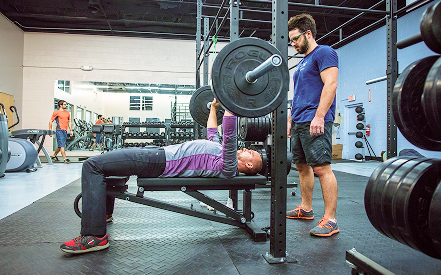 A person bench presses a barbell on a bench in a gym, assisted by a spotter. Both appear focused. The environment is filled with various gym equipment.