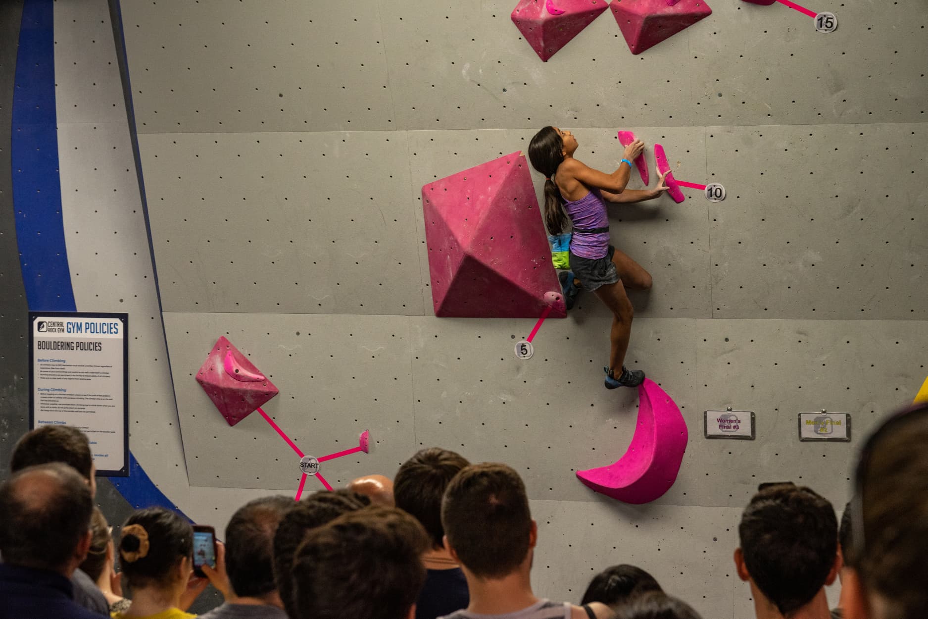 A teen girl climbs on a competition boulder. She holds onto crimpy sidepulls and has a drop knee.
