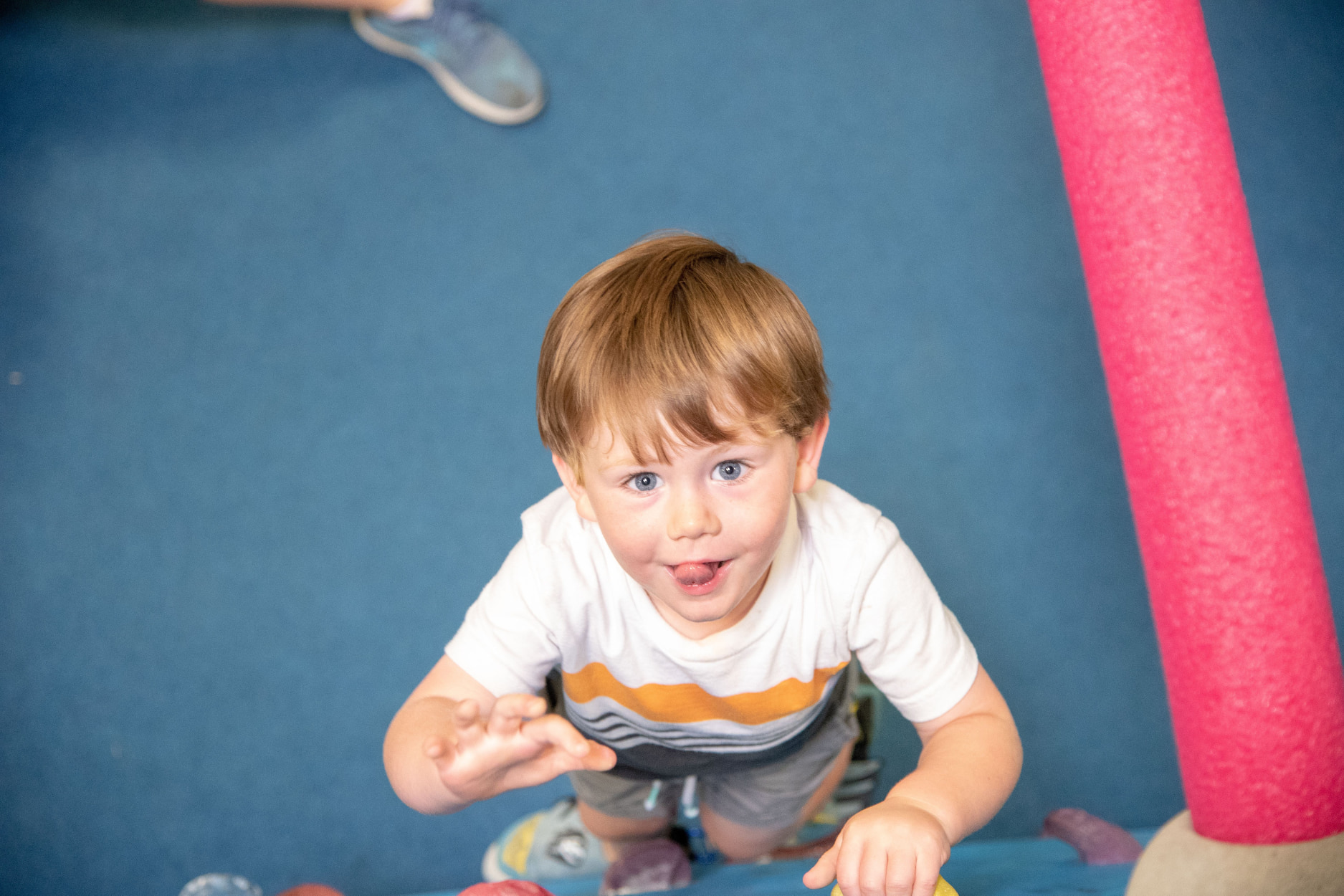A toddler climbs and sticks out his tongue in focus as he completes a bouldering challenge climbing under pool noodles.