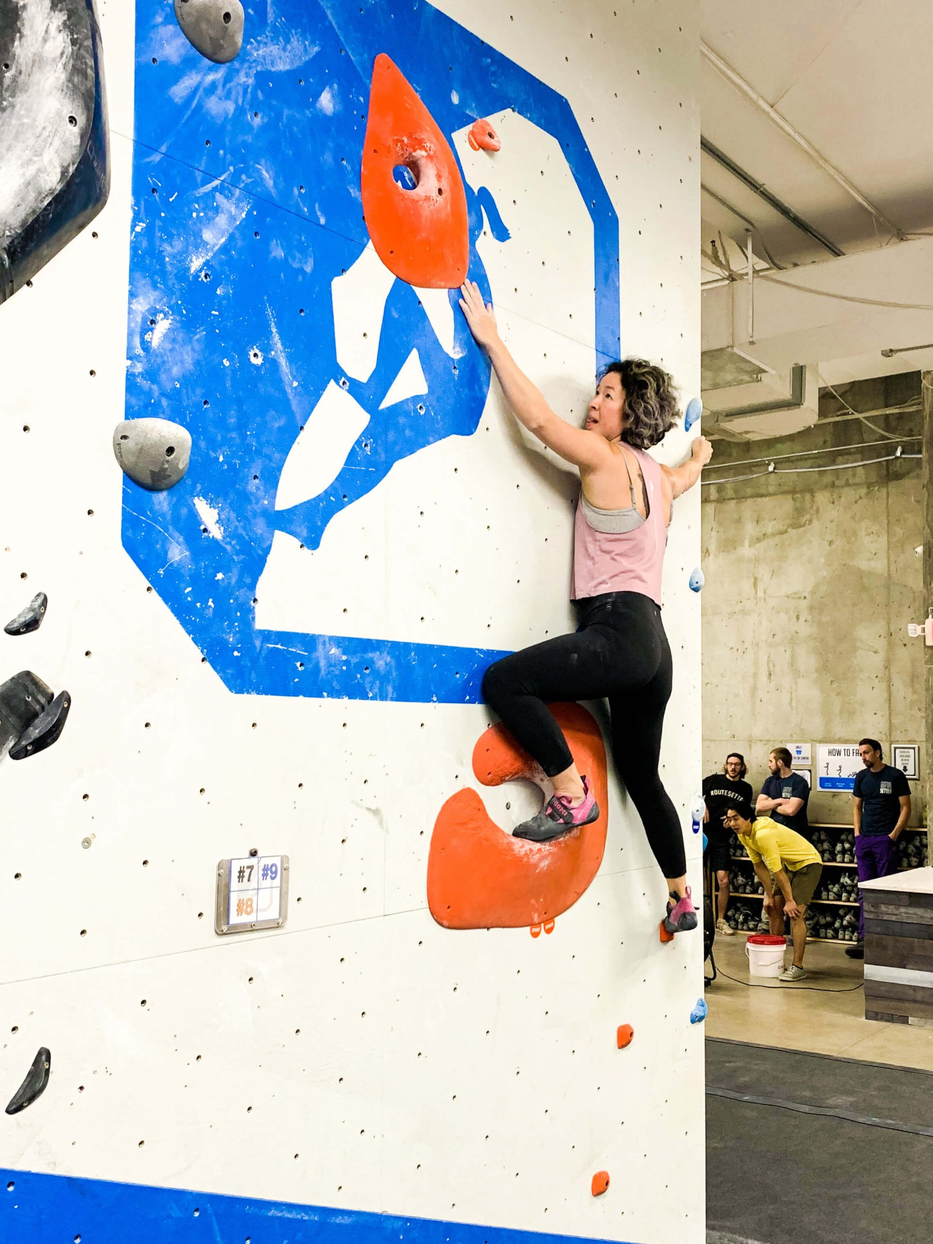 A woman with short hair is bouldering. She holds onto the arete with one hand and reaches for the next hold with the other.