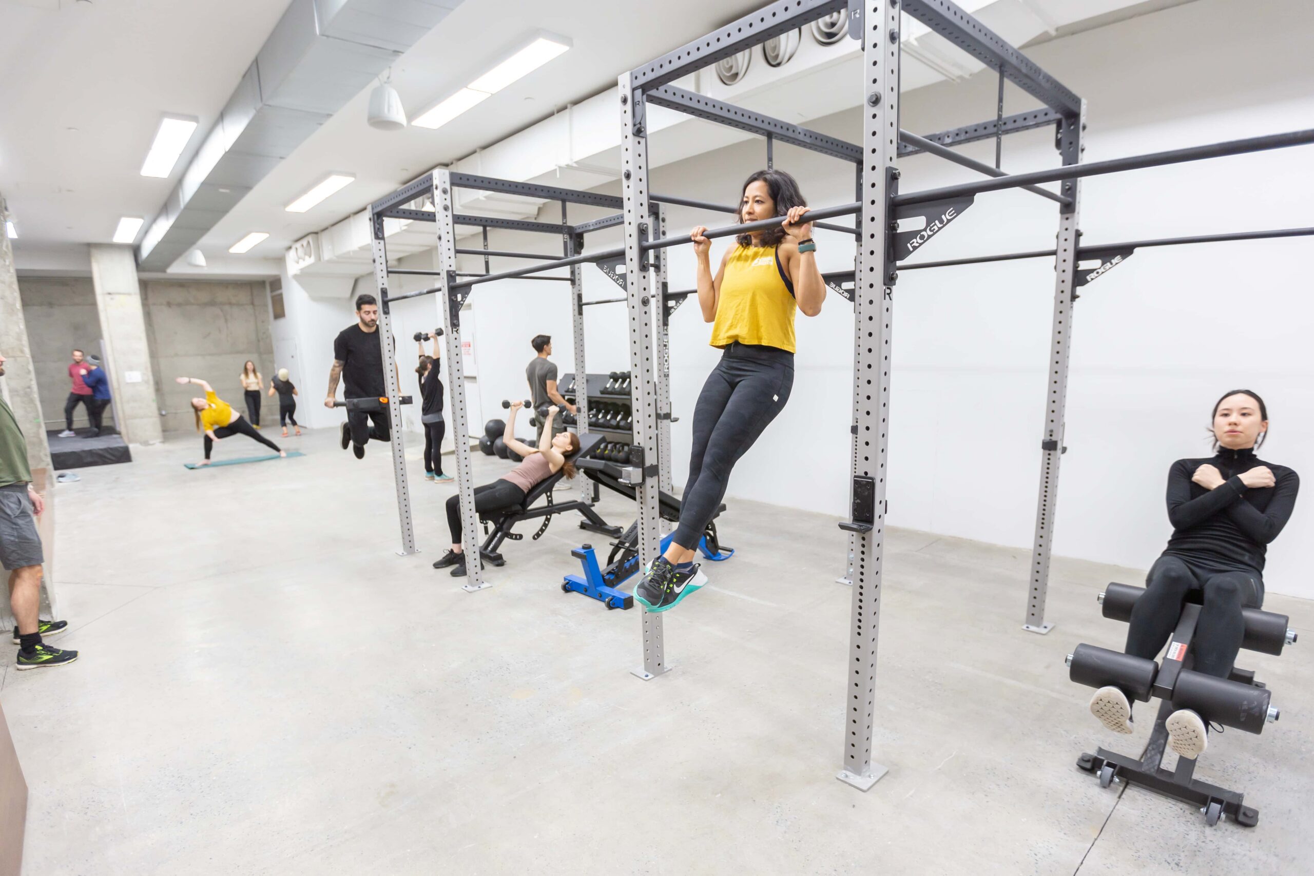 A woman trains pull ups while others work out in a gym area.