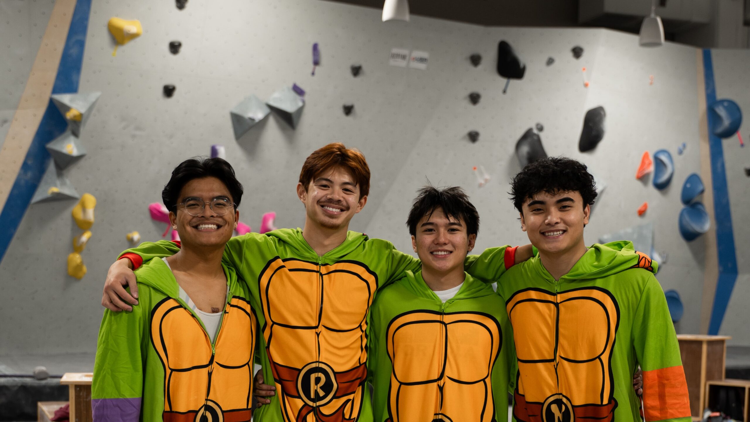 A group of young men pose together in front of a bouldering wall during an event at Central Rock Gym Citrus Park. They are dressed up in Ninja Turtle onesies.
