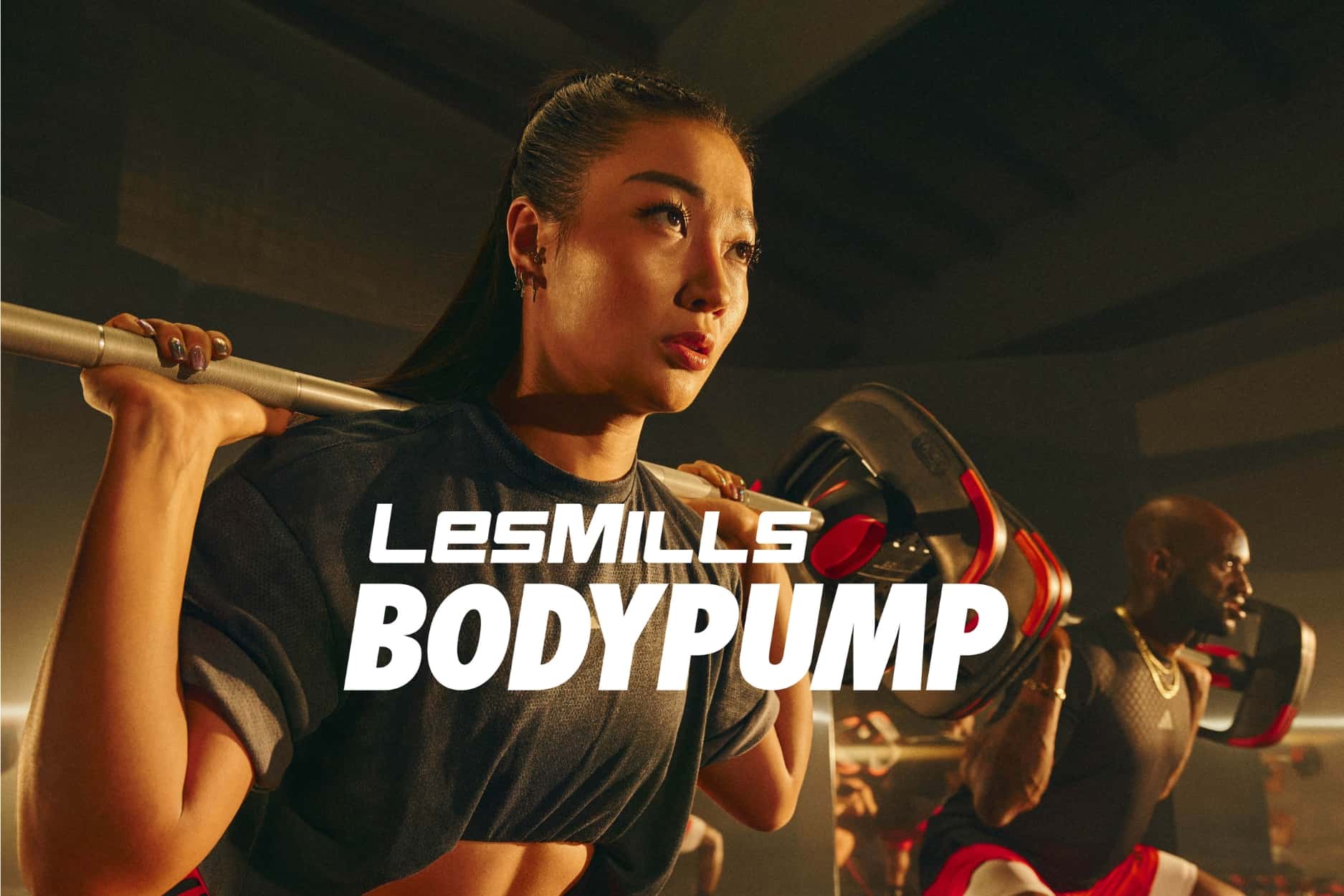 A woman squats with a barbell on her back as others workout in the background. She is focused on her breath. Text overlay reads LesMills Bodypump.