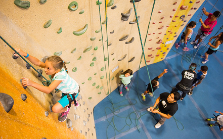 A young girl confidently climbs an indoor rock wall, watched by a group of children below. Instructors assist, creating a lively and encouraging atmosphere.