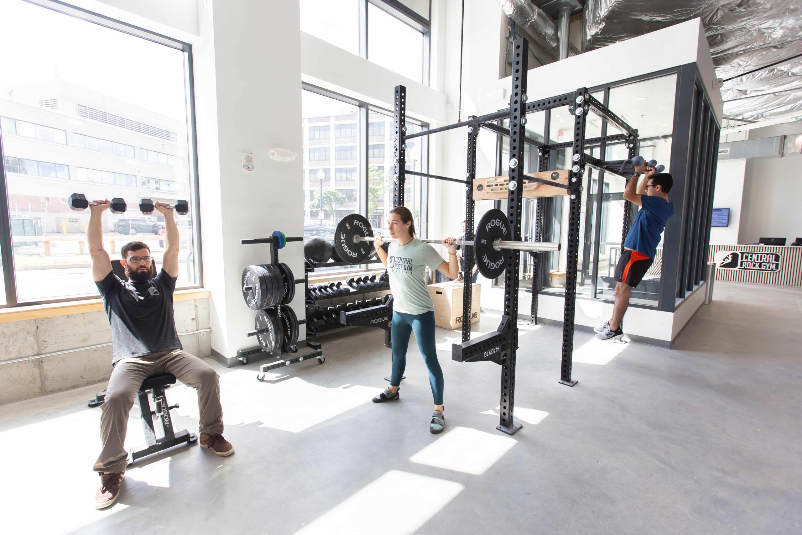 Members work out in the fitness area at Central Rock Gym Fenway — a man presses dumbbells overhead, a woman squats a barbell, and another member does pull-ups on the rig