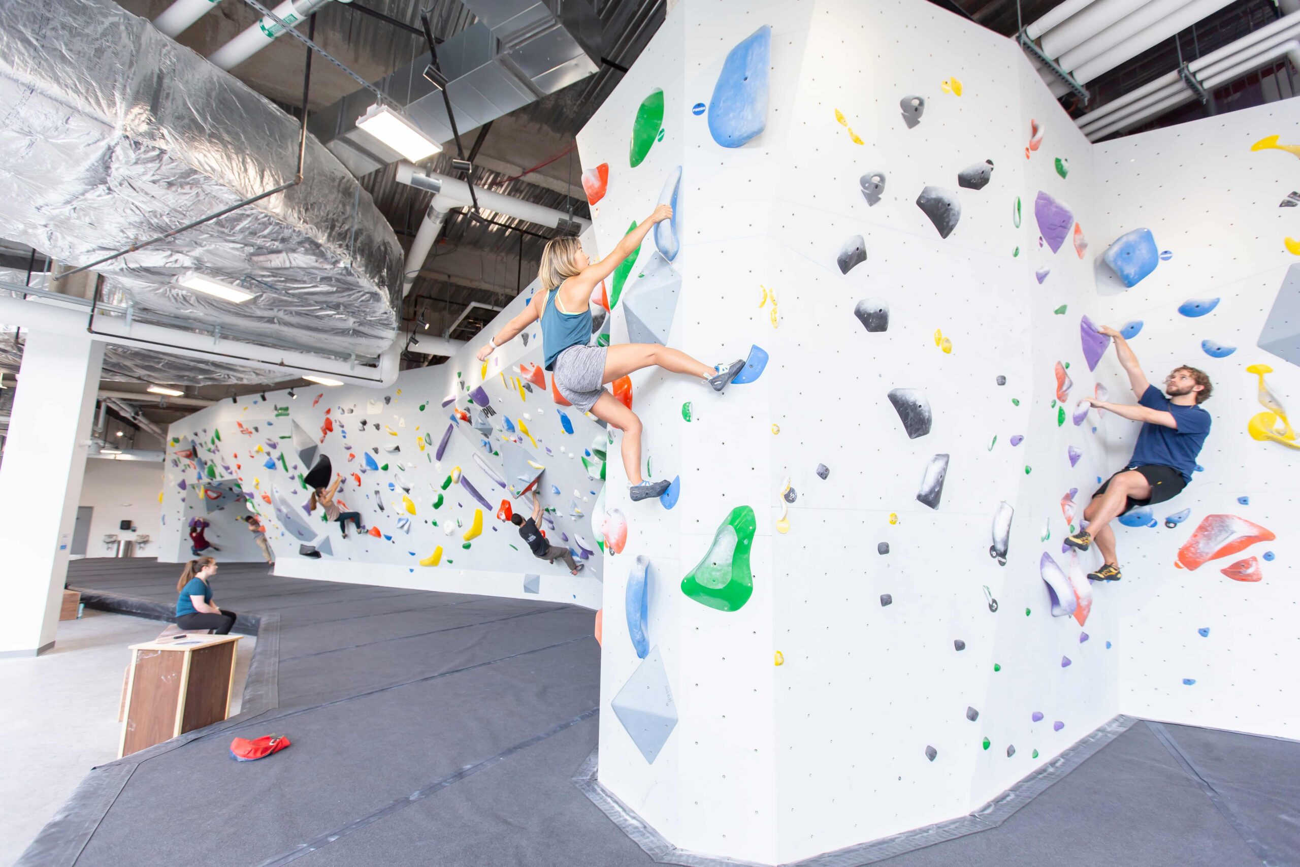 A woman leans into a sidepull on the bouldering wall at Central Rock Gym Fenway while another climber works a route beside her