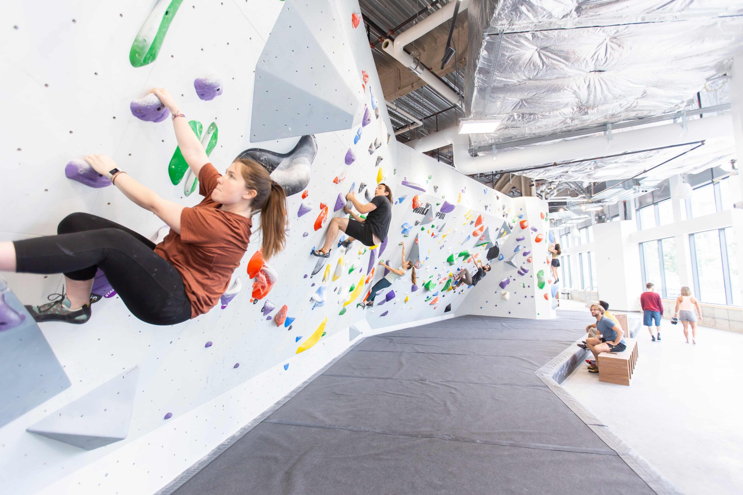 A woman sends a bouldering problem in the foreground at Central Rock Gym Fenway while several other climbers work routes on the sweeping white walls behind her, with crash pads on the floor and large windows flooding the gym with natural light