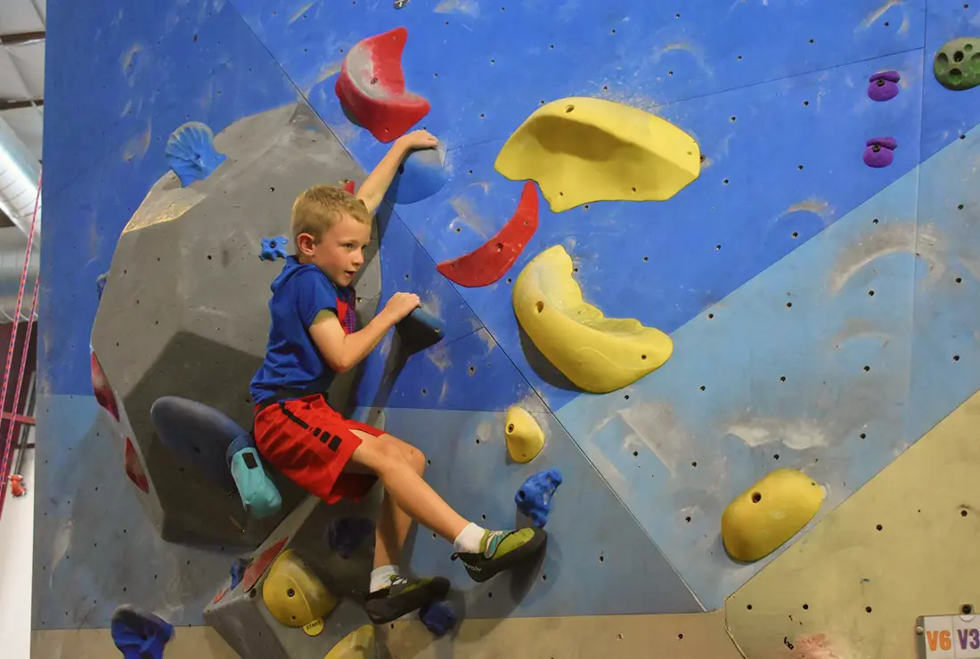 A young kid in a blue shirt and red shorts boulders on the blue wall at Central Rock Gym