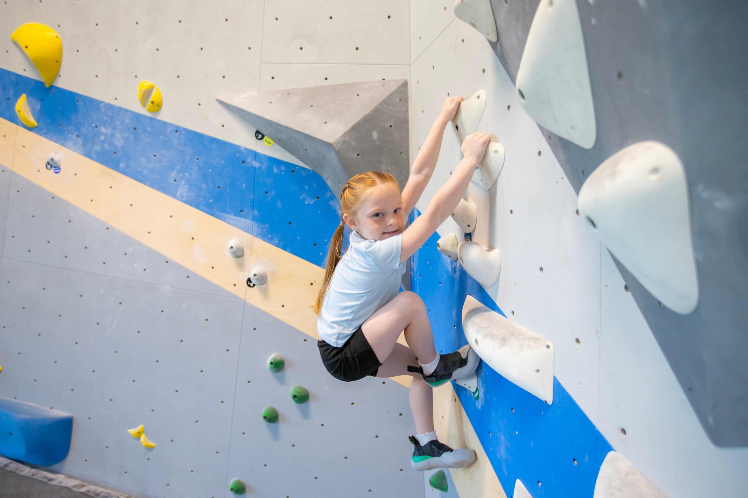 A young girl with pigtail braids boulders and looks sideways at the camera.