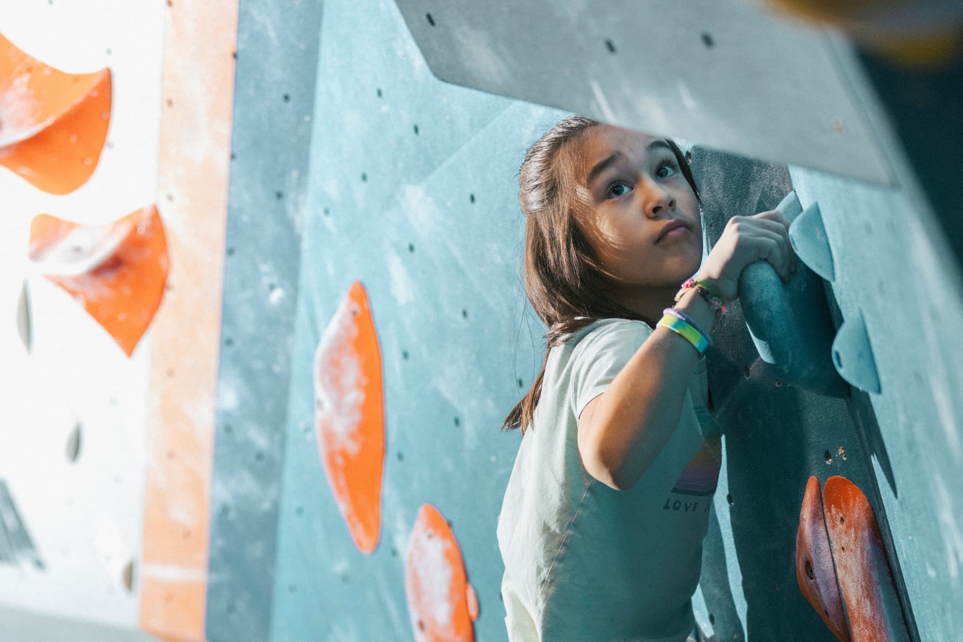 A young girl holds onto a jug and looks at the next climbing hold on the bouldering wall.