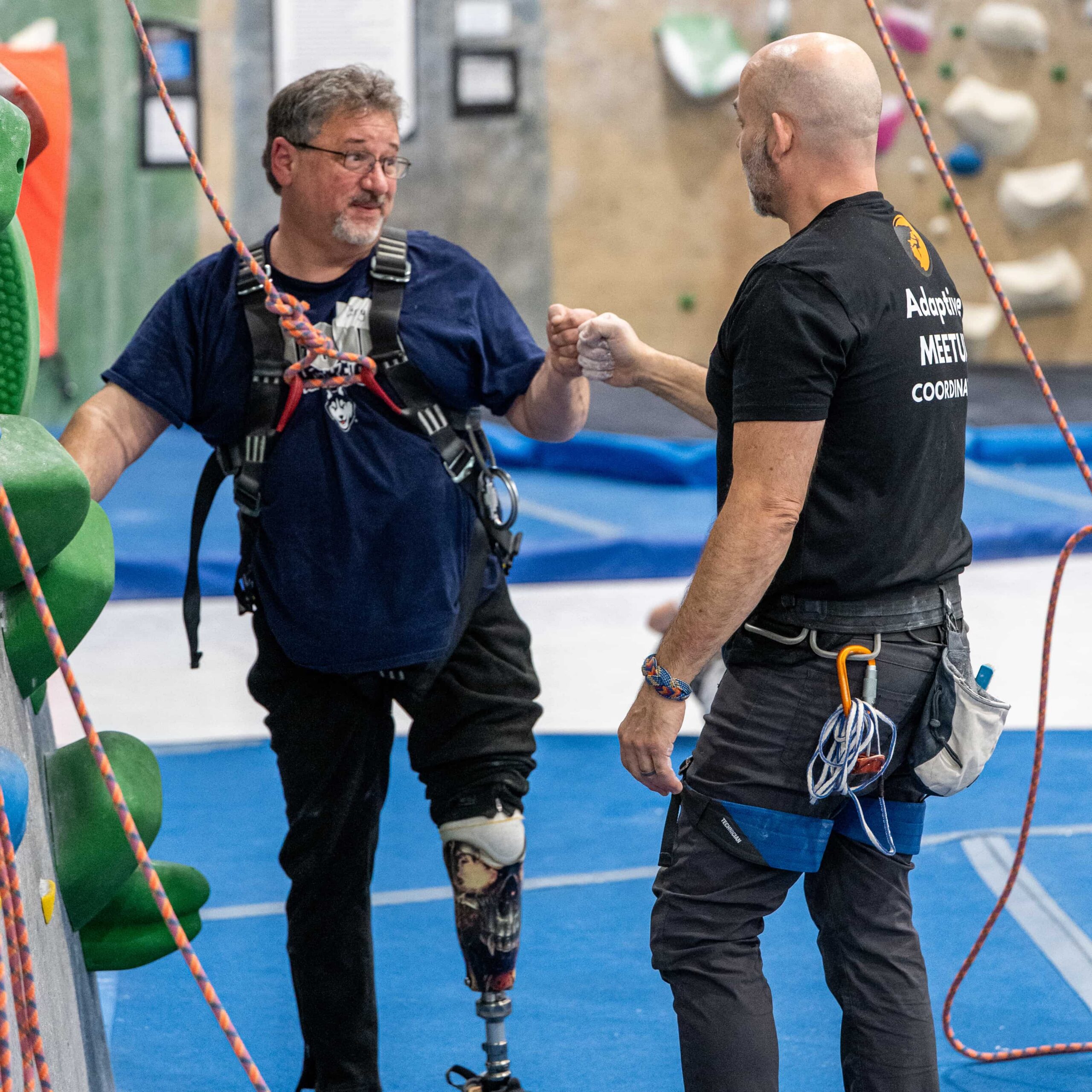 A climber with a chest harness and prosthetic leg fist bumps his belayer after completing a climb.
