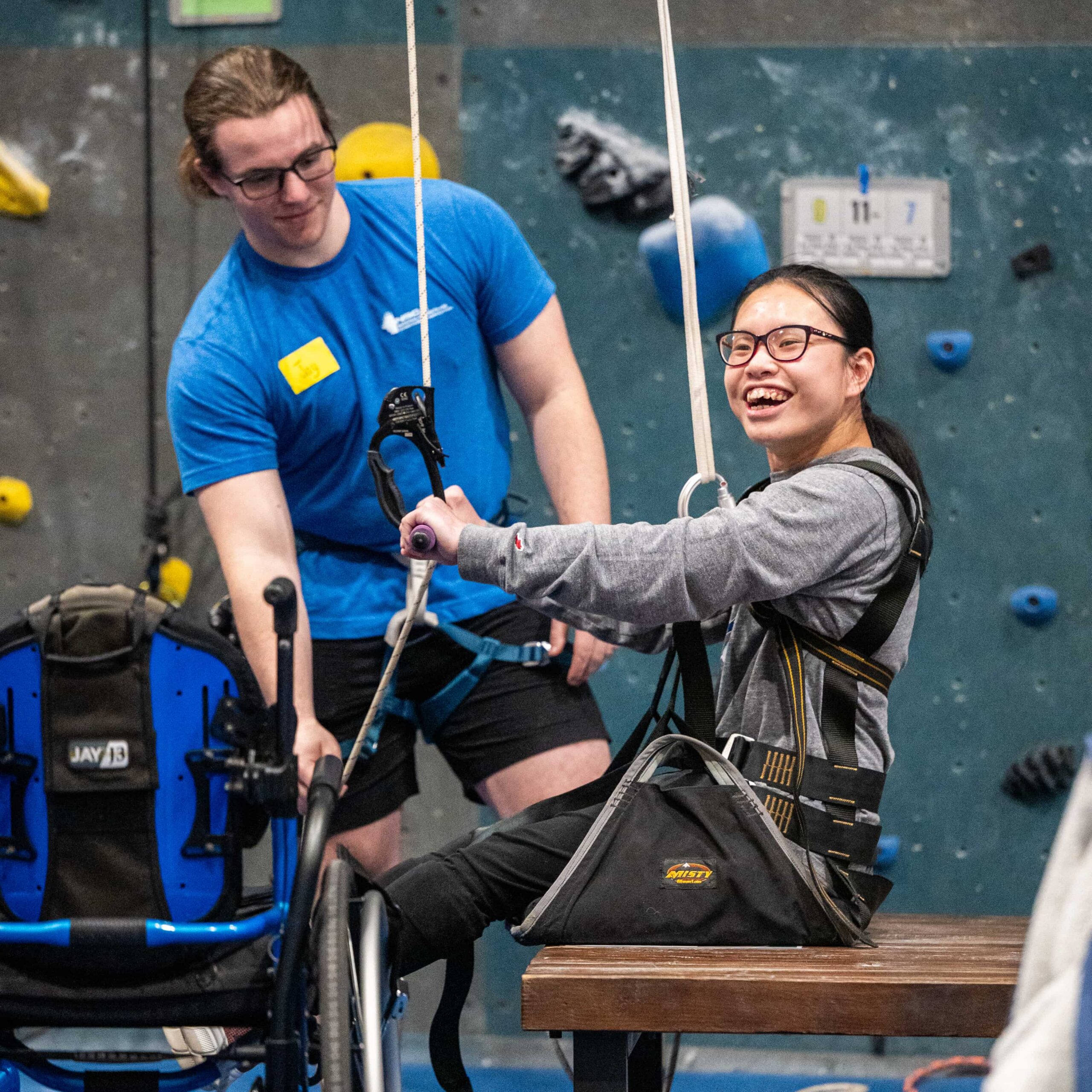 An adaptive climber beams with a smile as she start climbing using a seat harness and pull up bar.
