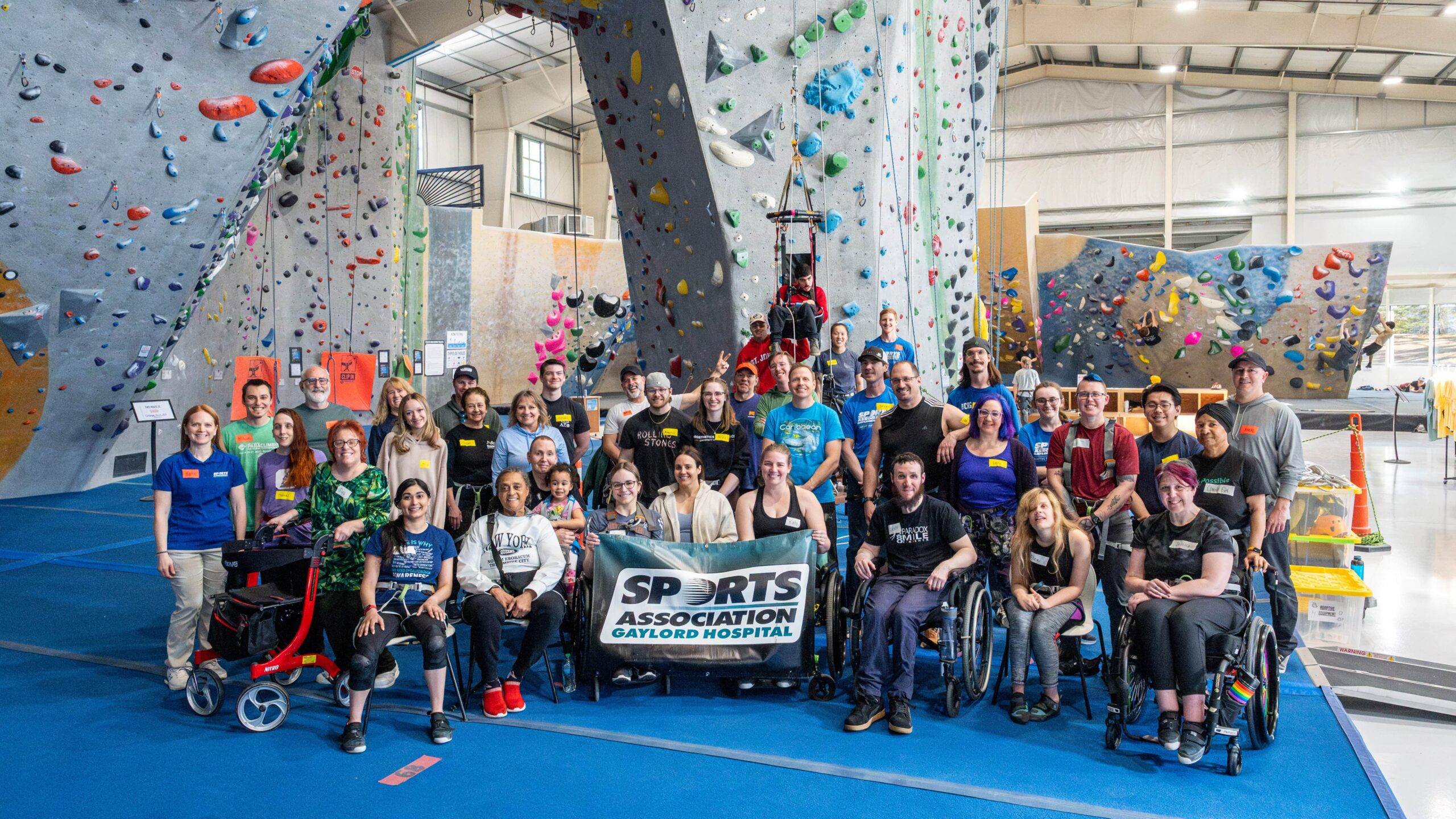 Adaptive climbers posing for a photo in front of a tall indoor climbing wall.