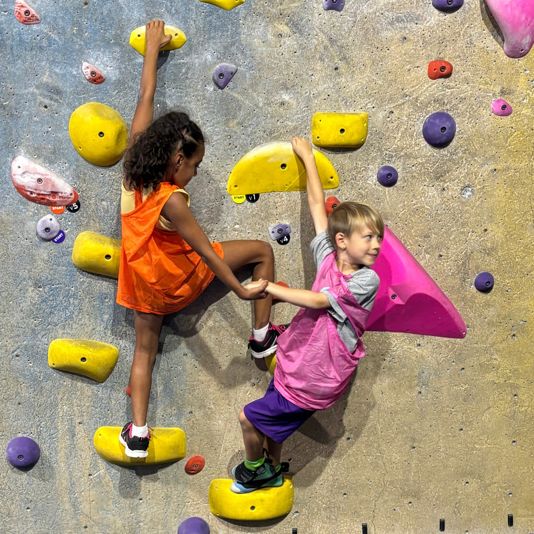 Two kids holding hands while climbing on a bouldering wall.