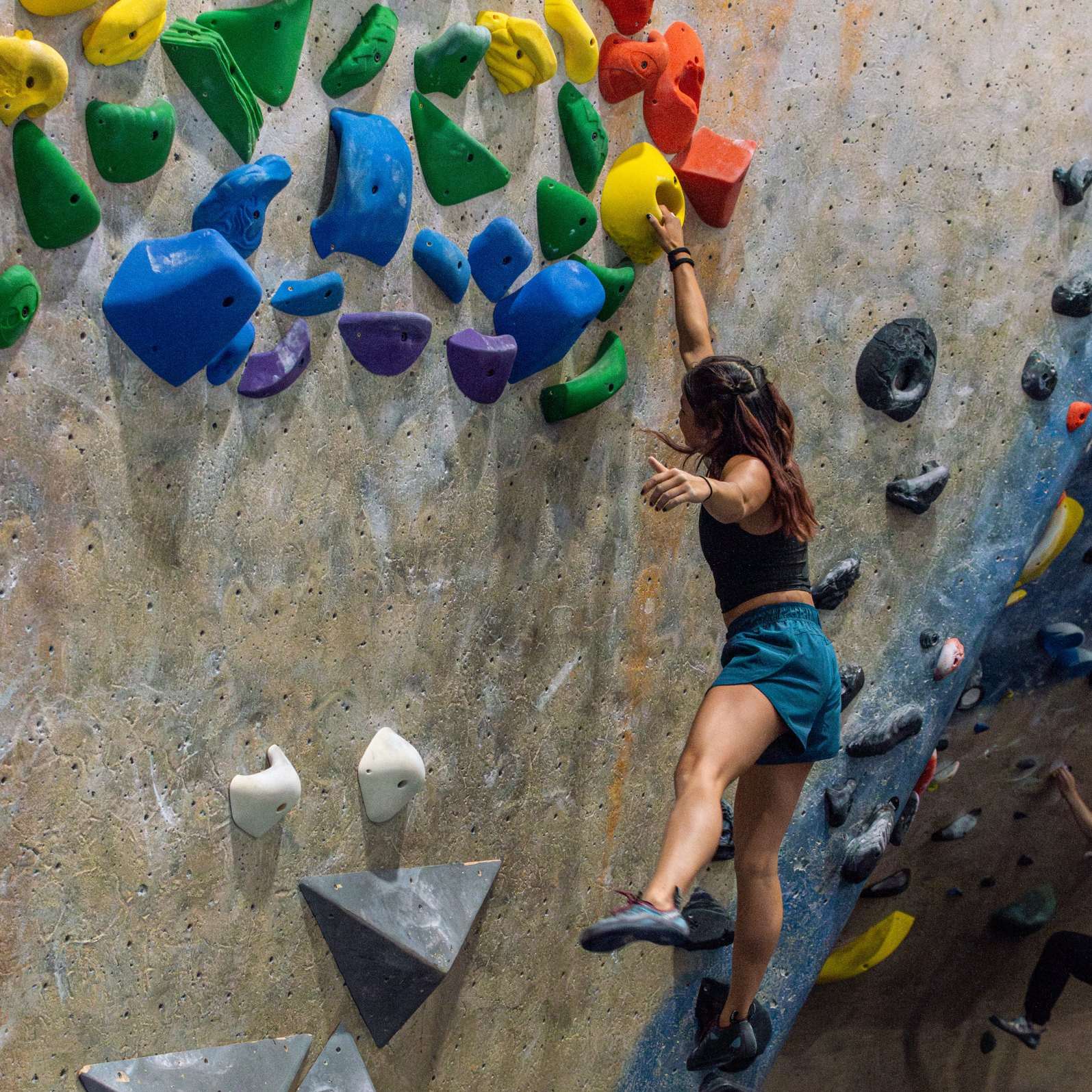 A climber bouldering on an indoor climbing wall, doing a dynamic move. There are rainbow colored holds on the wall.
