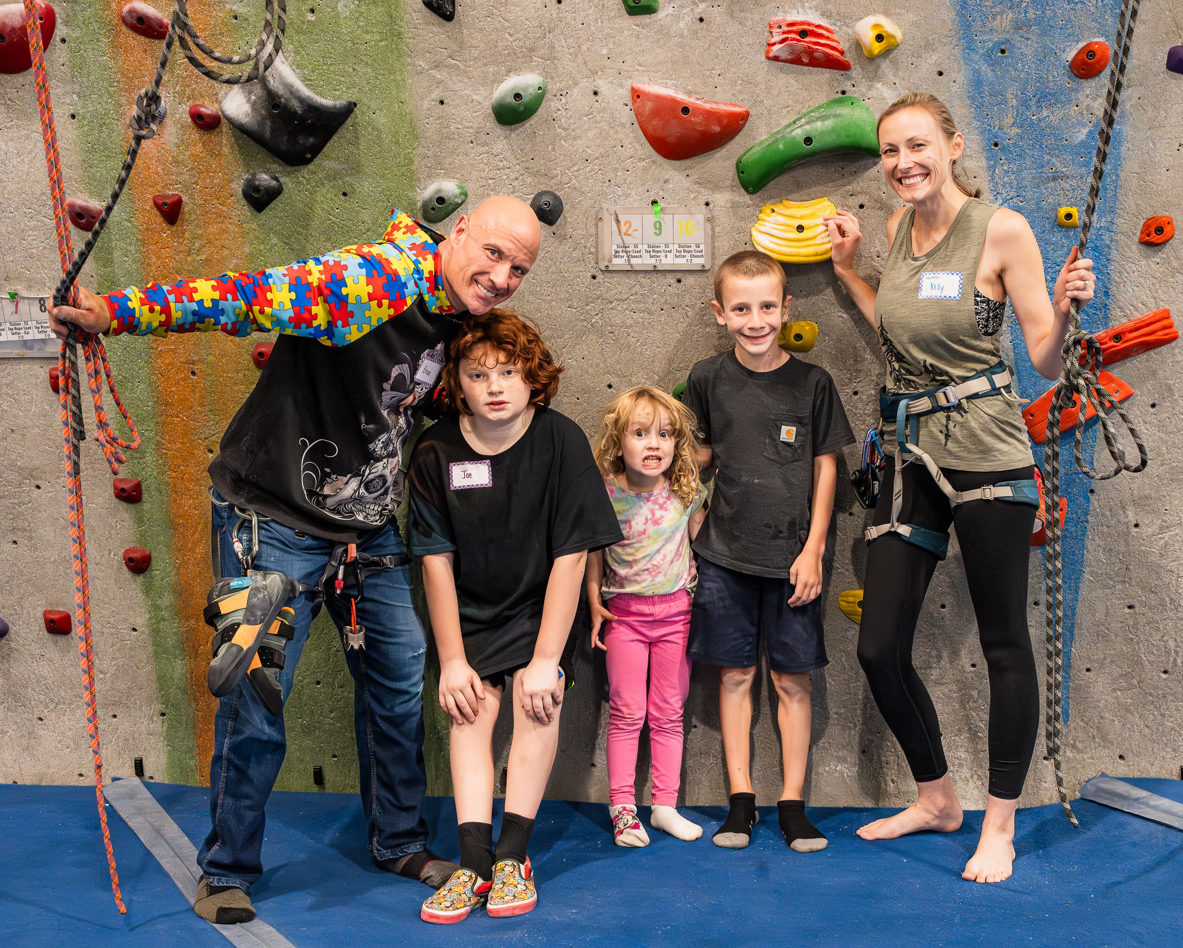 A family with two young sons and a young daughter poses in front of a climbing wall. The dad has on a shirt with puzzle pieces on the sleeves and they are all wearing name tags.