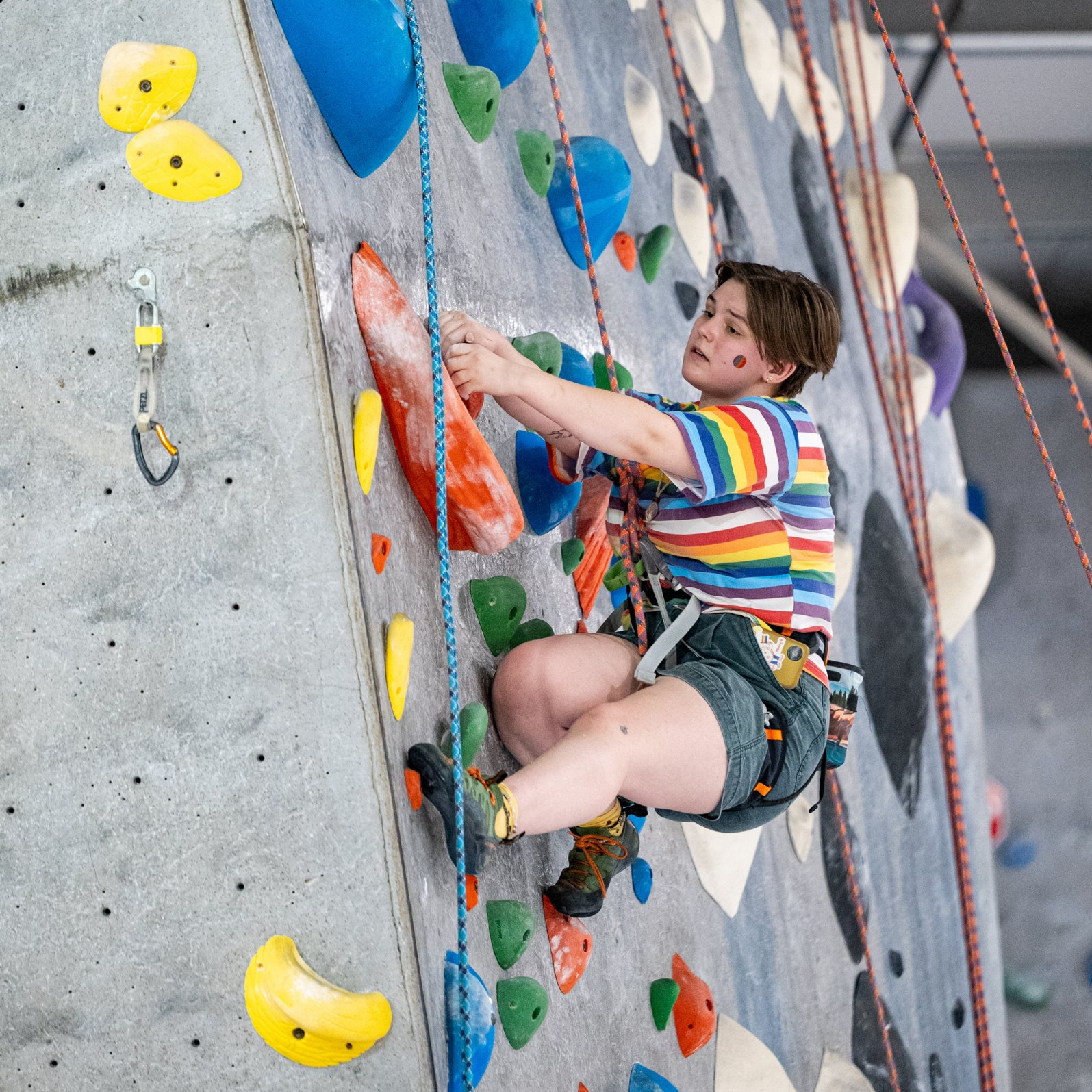 A climber wearing a colorful shirt while rope climbing. Climber is reaching with their left hand.