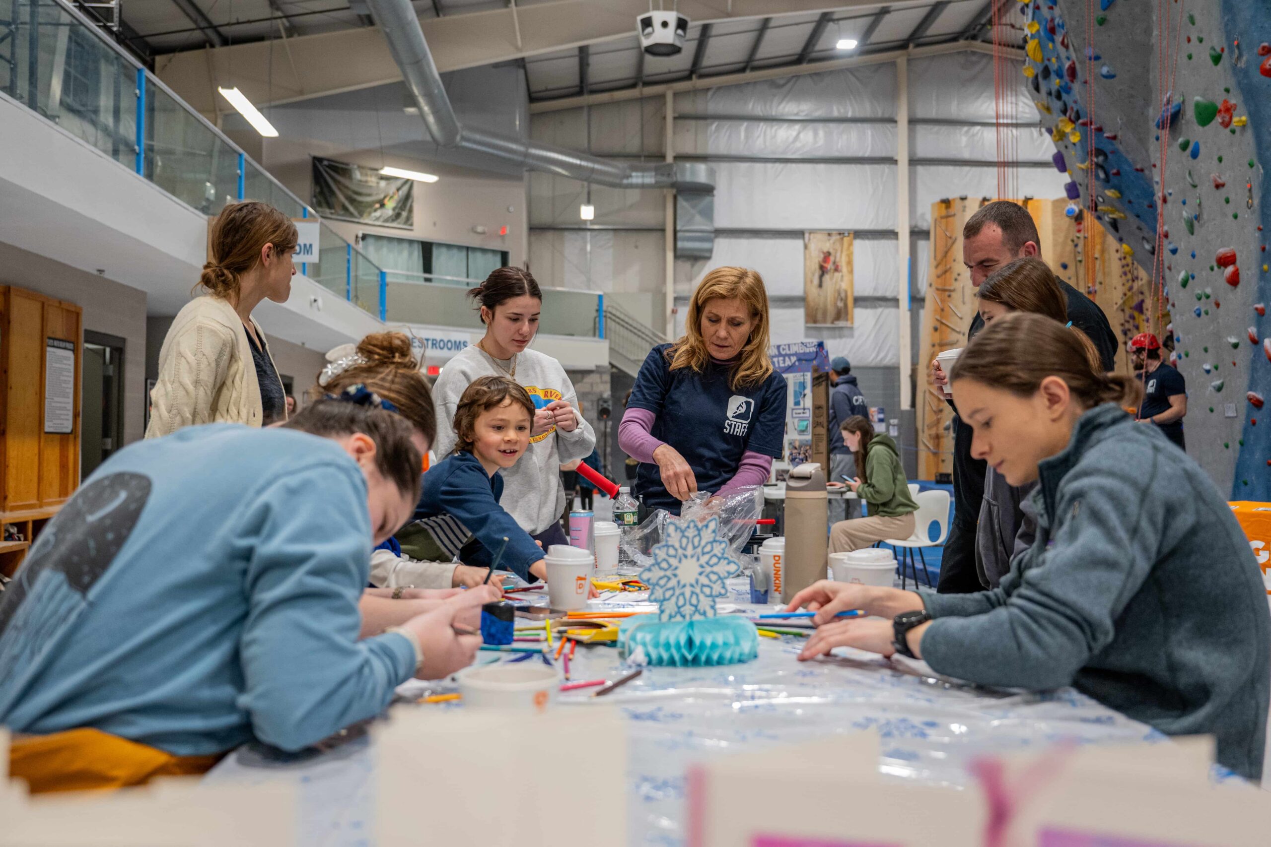 A woman wearing a CRG staff shirt stands at a table full of people working on making winter themed crafts.