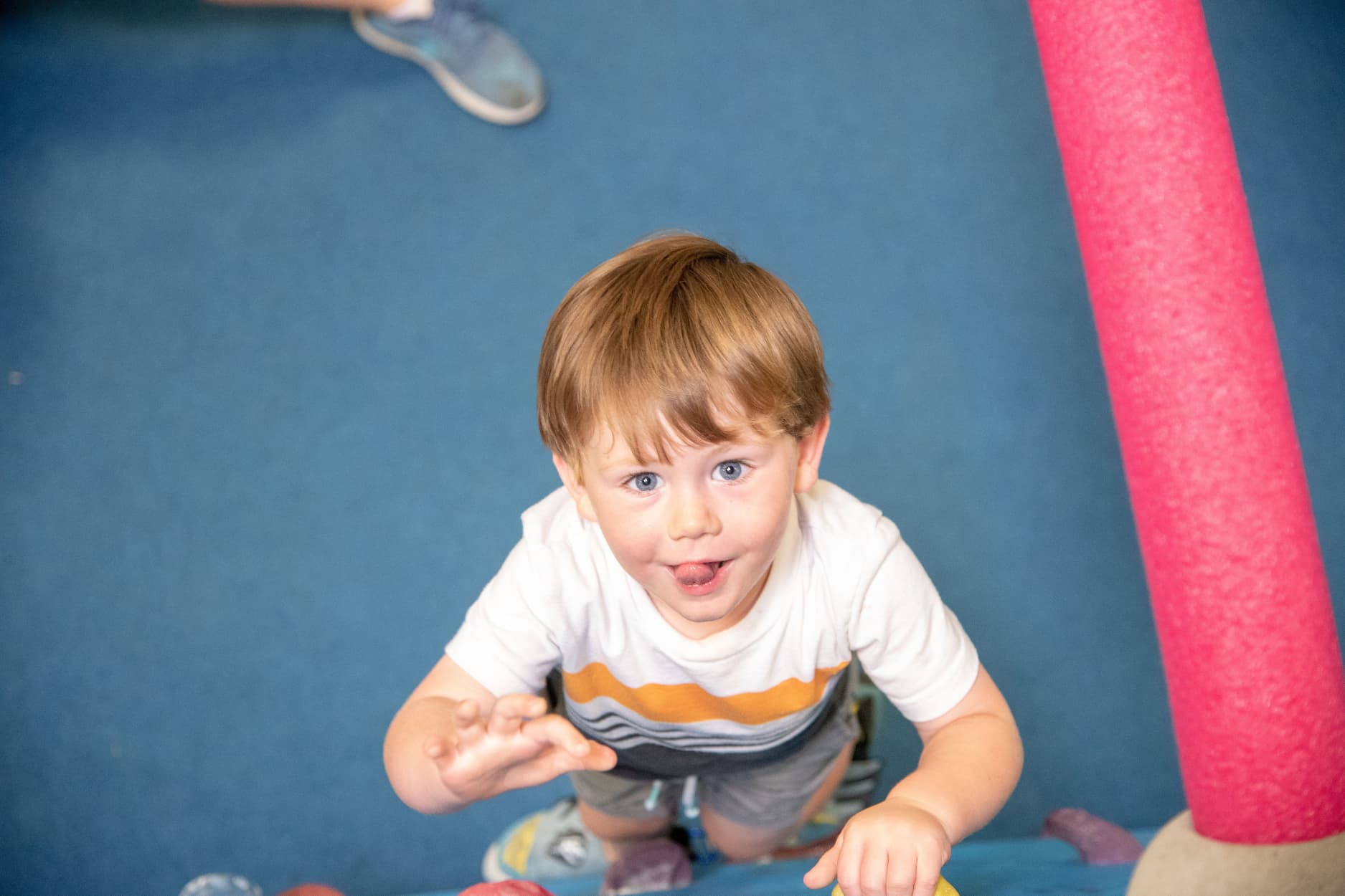 A very young boy sticks his tongue out as he focuses on hanging onto a bouldering wall just off the ground.