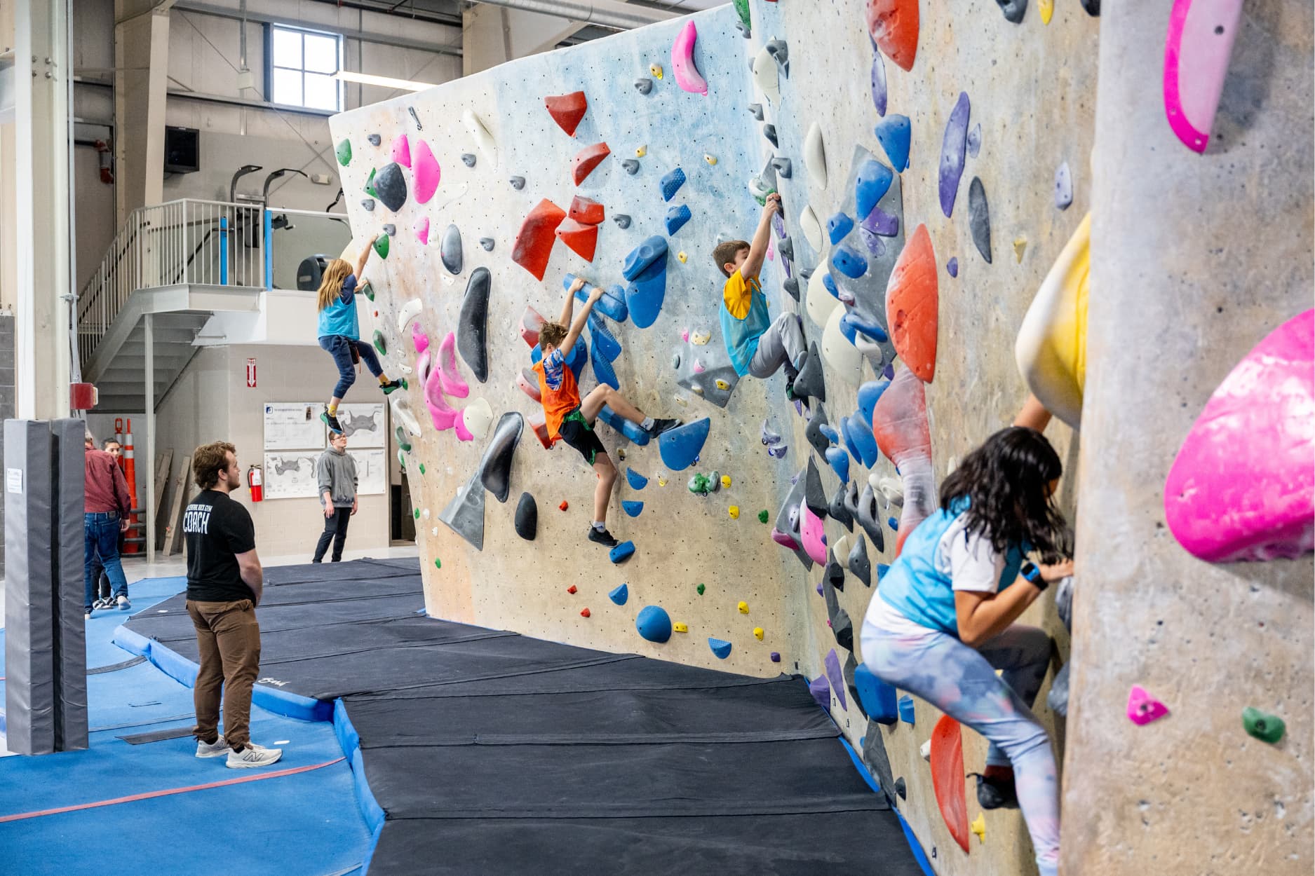 A CRG coach stands and watches kids boulder during a youth program. They are wearing colorful jerseys and completing a bouldering challenge.