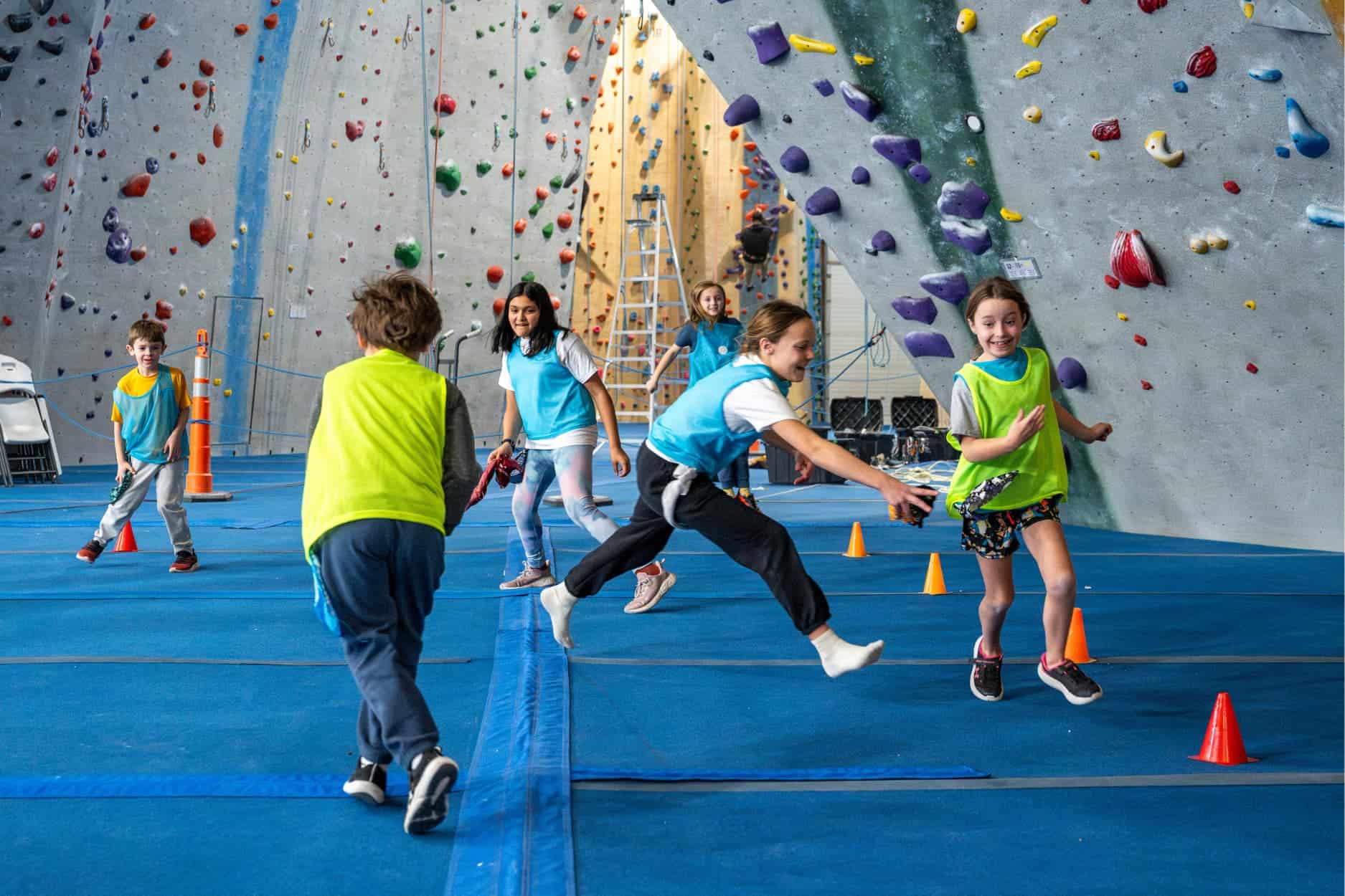 A group of kids are wearing two different colors of jerseys that divide them into teams. They are running around chasing each other at Central Rock Gym playing a game where they try to grab a bandana that is tucked into each other's pockets and hanging out like a tail.