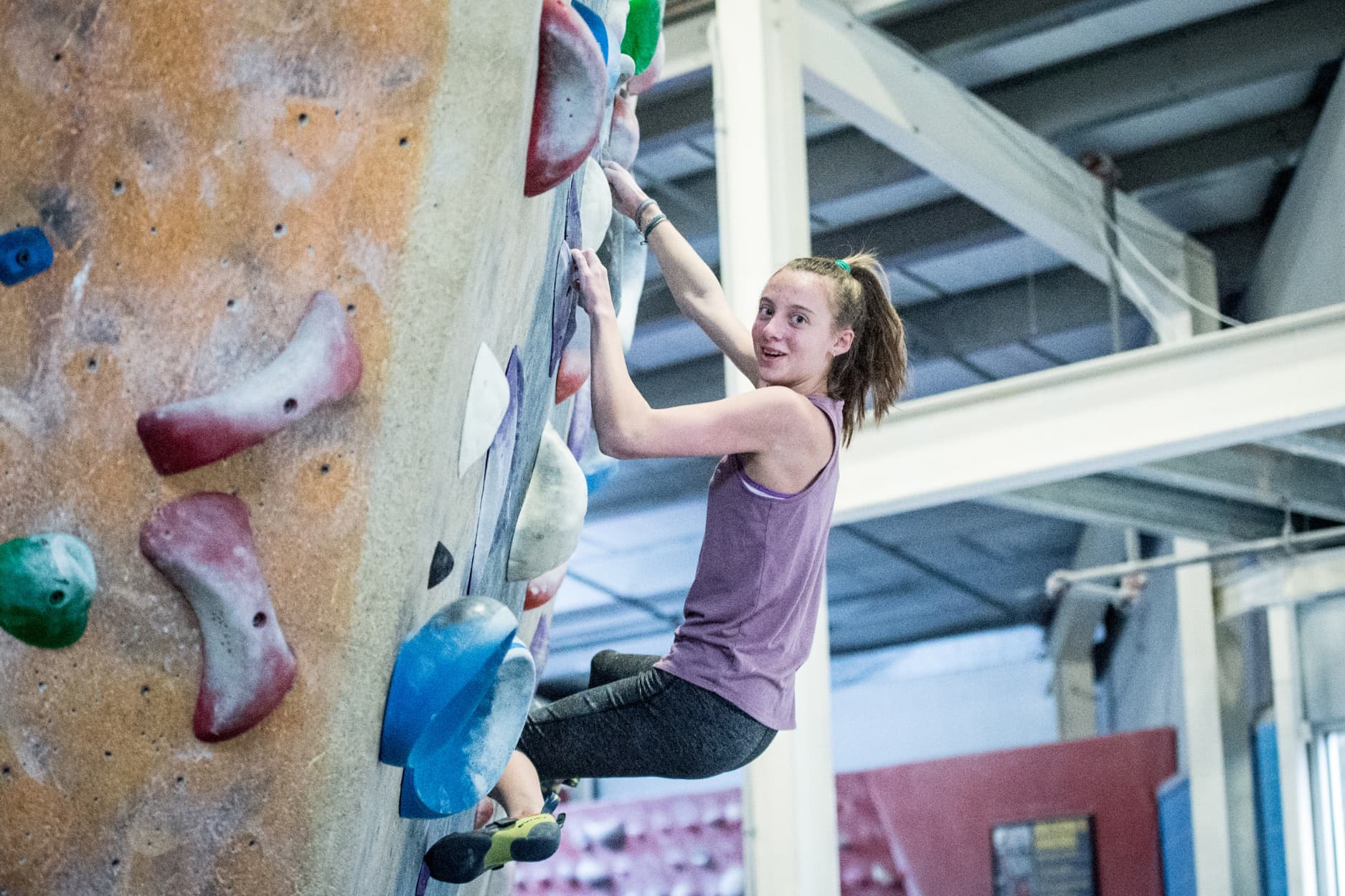 A teen girl smiles at the camera while climbing a crimpy boulder.