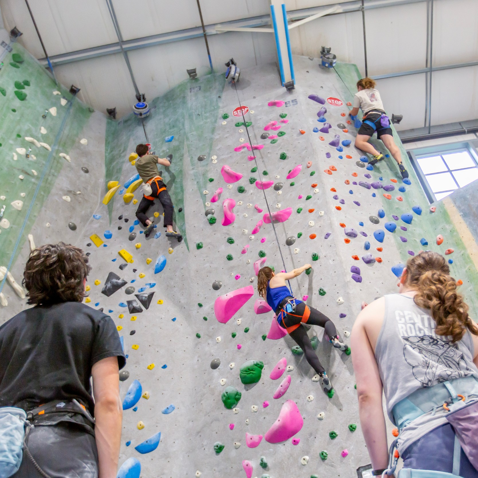 Three people climbing on auto belays at an indoor rock climbing facility. There are two people standing in the foreground watching the climbers.