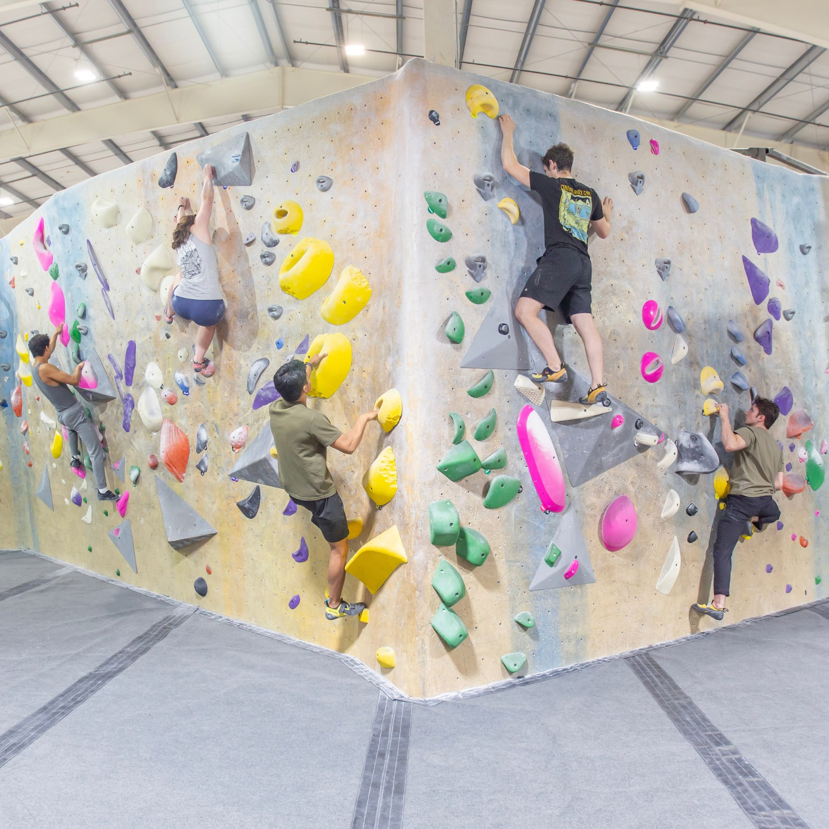 Five climbers bouldering at an indoor rock climbing facility. The wall features colorful climbing holds and a gray pad underneath.