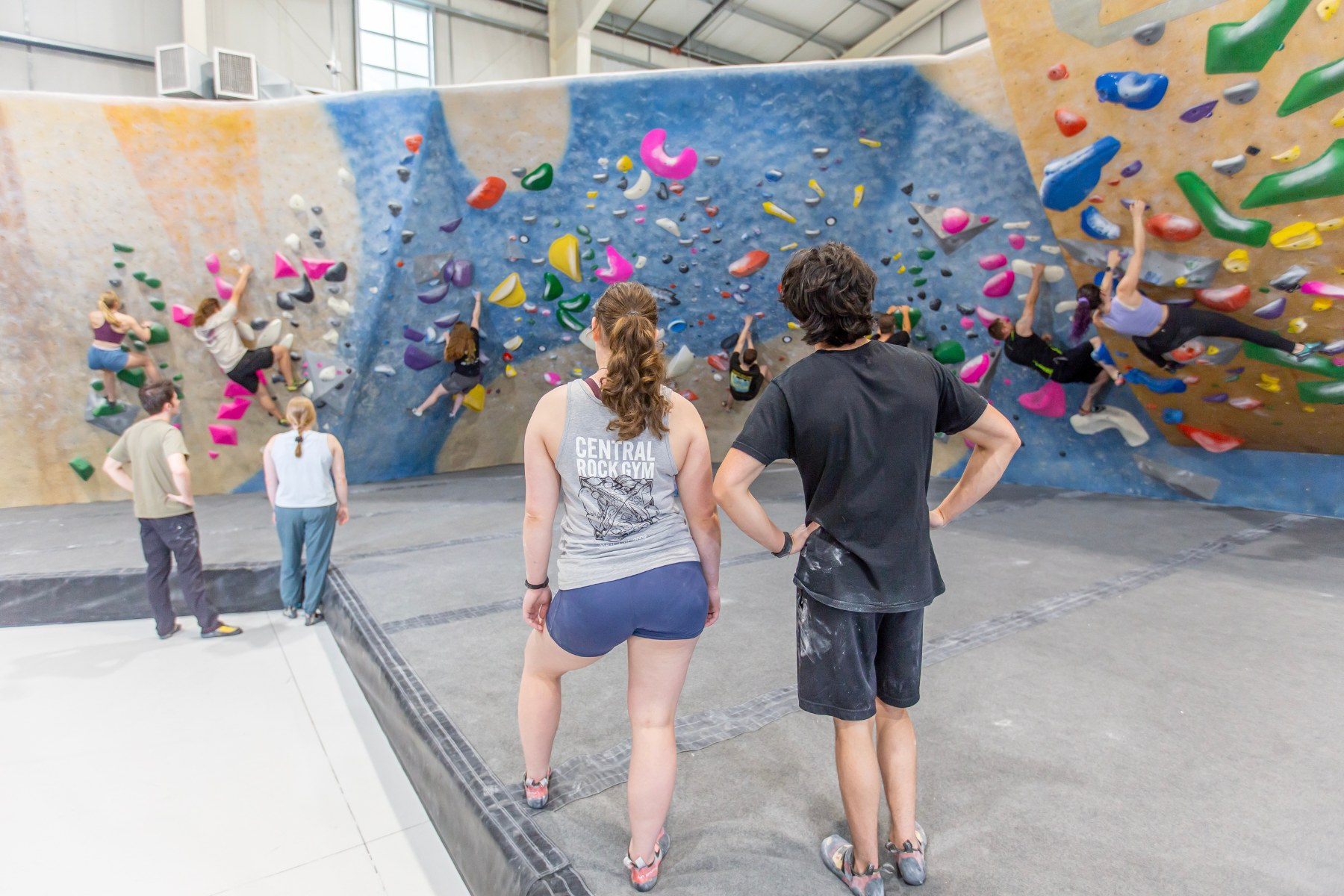 Several climbers are bouldering on an indoor climbing wall while others watch. The wall features colorful climbing holds and a gray pad underneath.