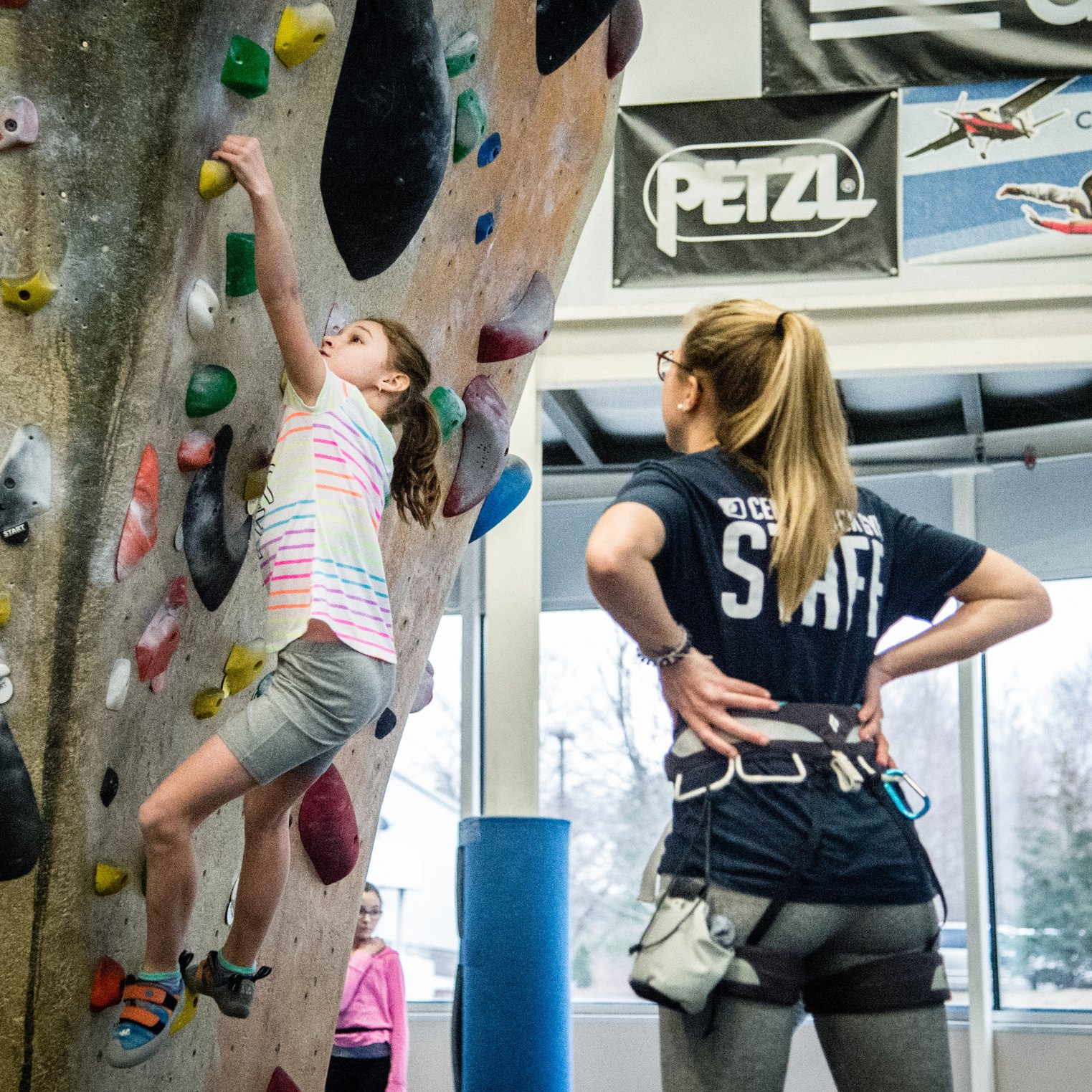 A CRG coach stands with her hands on her hips and watches a young girl on the youth team bouldering.