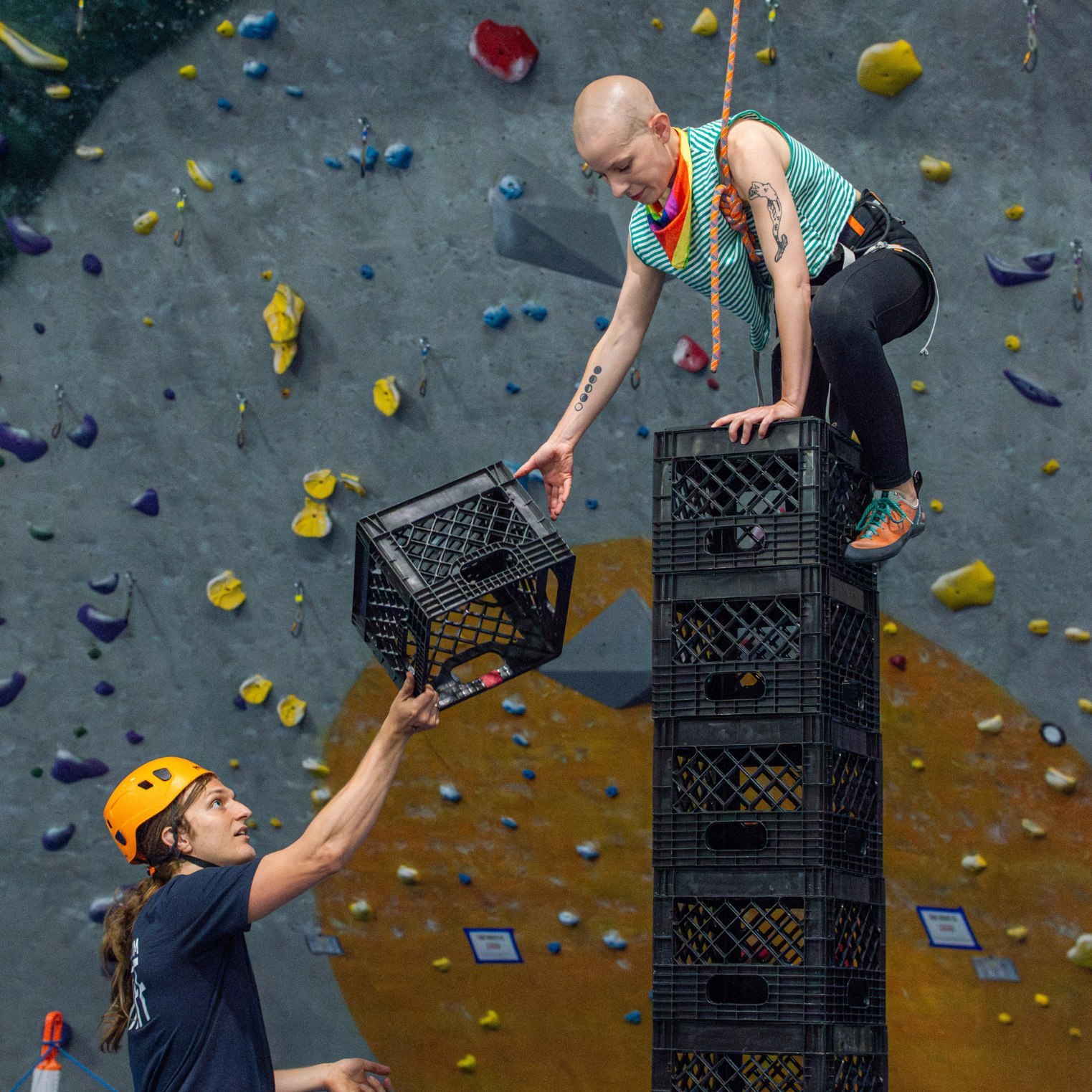 A woman climbing up a stack of milk crates attached to a top rope. A staff member is handing her another milk crate.