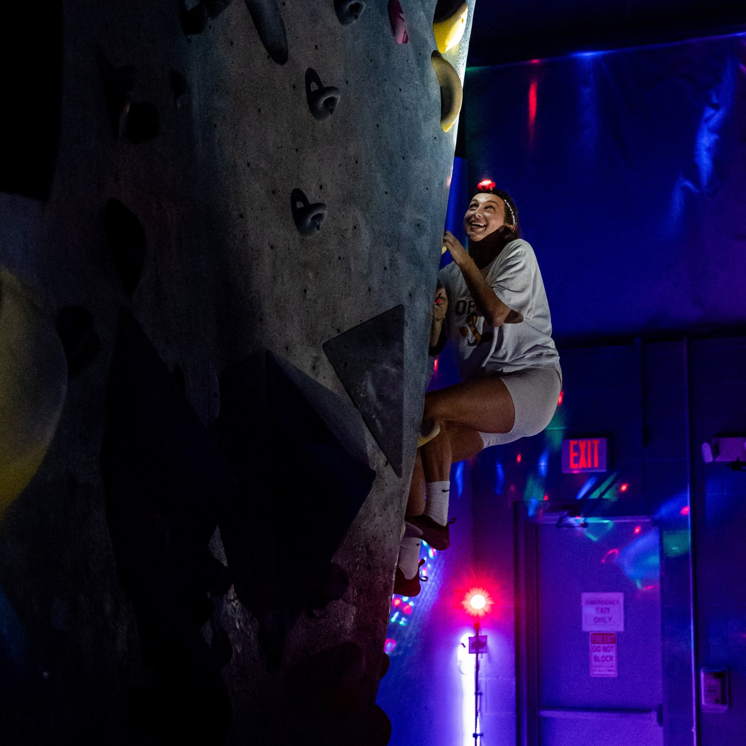 A woman climbing on an indoor bouldering wall wearing a headlamp. There are neon lights in the background.