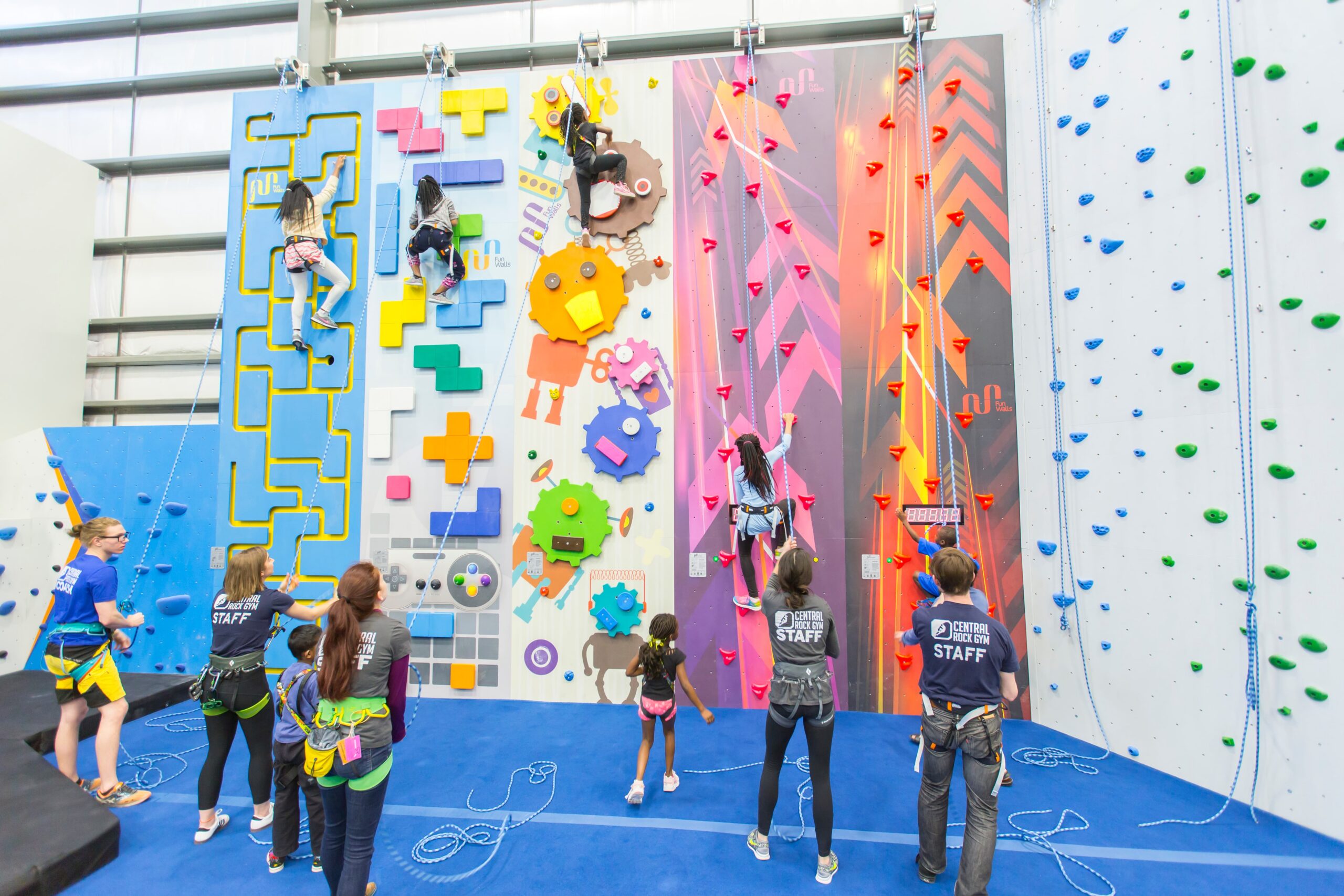A big group of kids on a tall climbing wall. They're being watched and belayed by mutlple staff members.