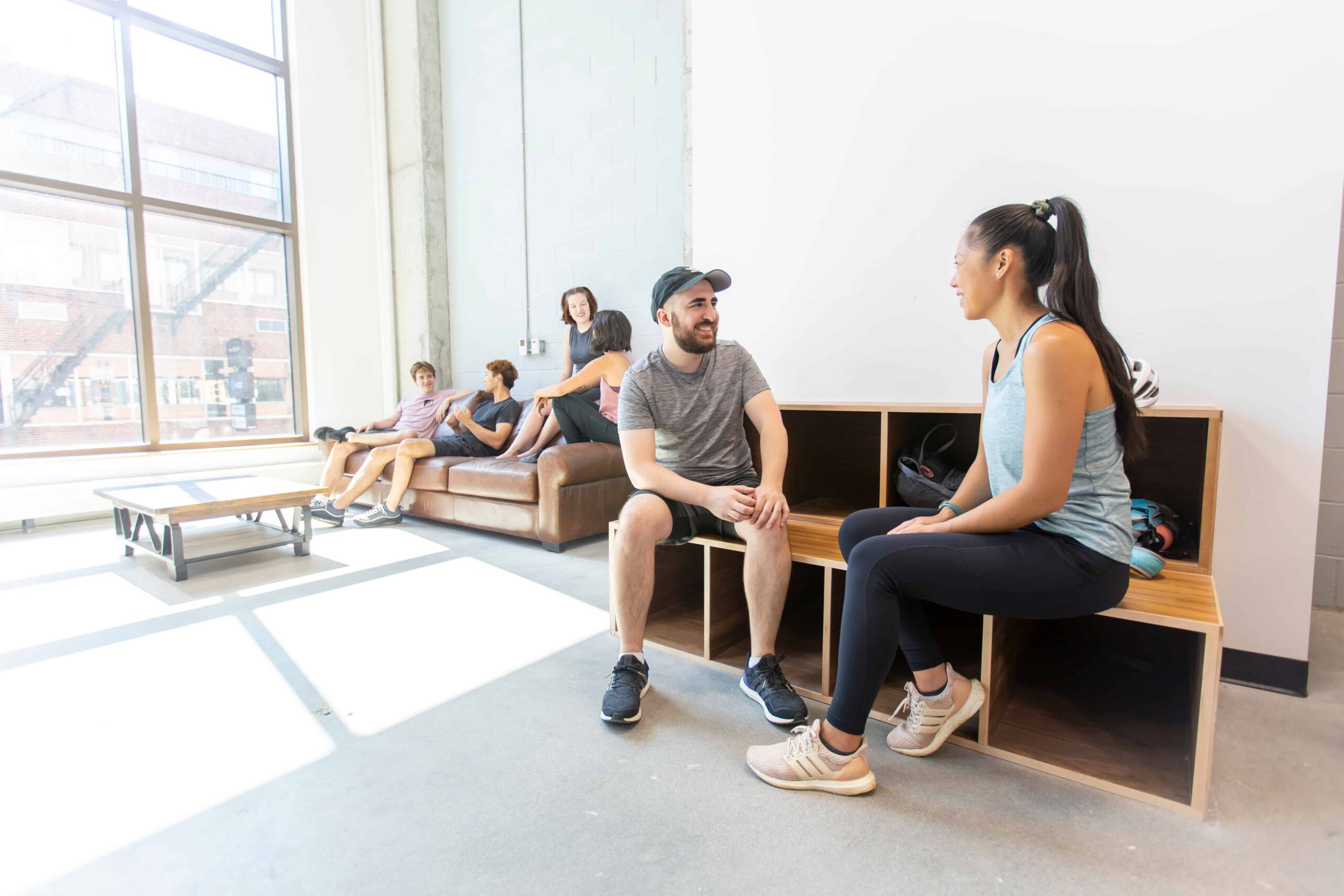 A man and woman sit on the benches and chat. In the background a group lounges on the couch.