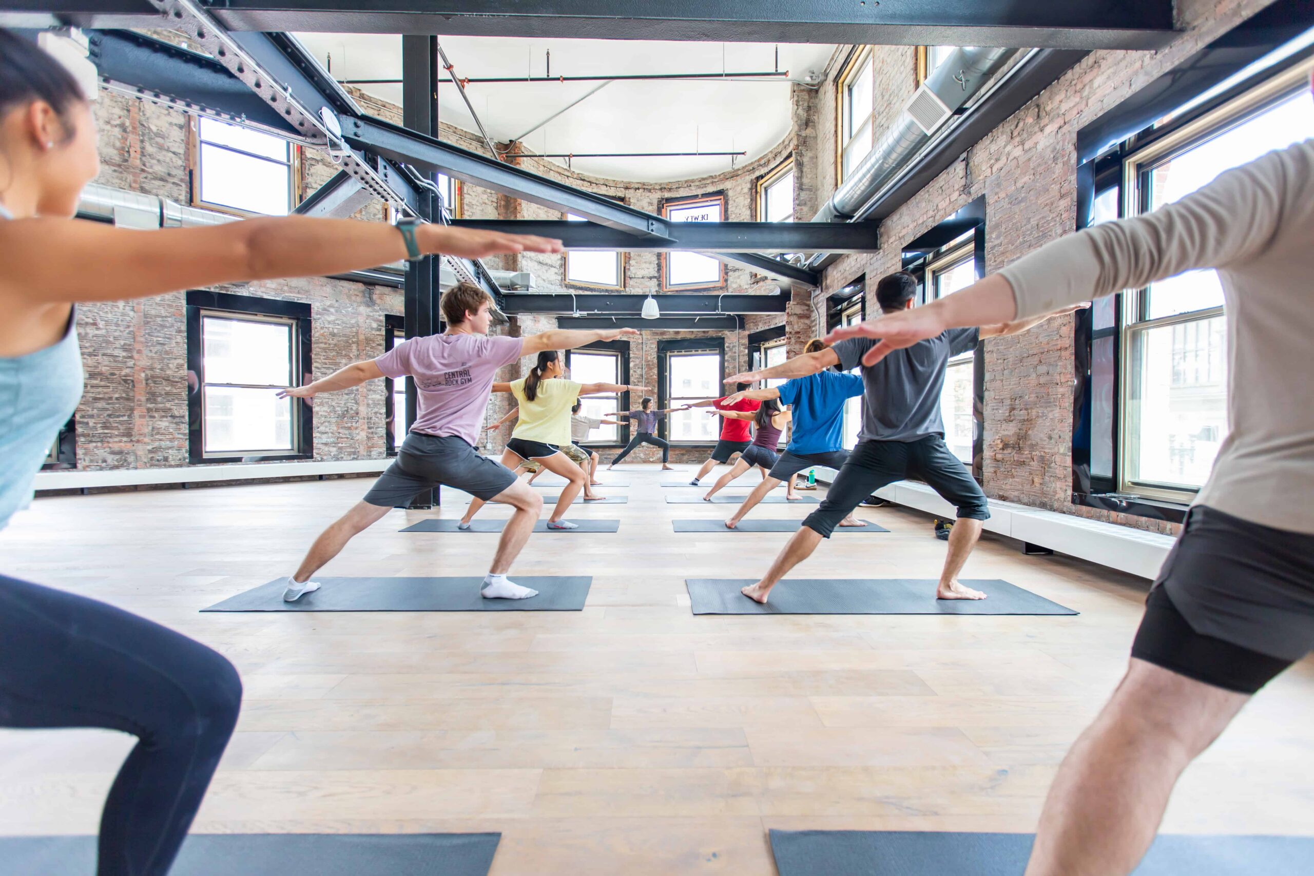 A yoga class doing Warrior 2 in a brick room with lots of natural light.