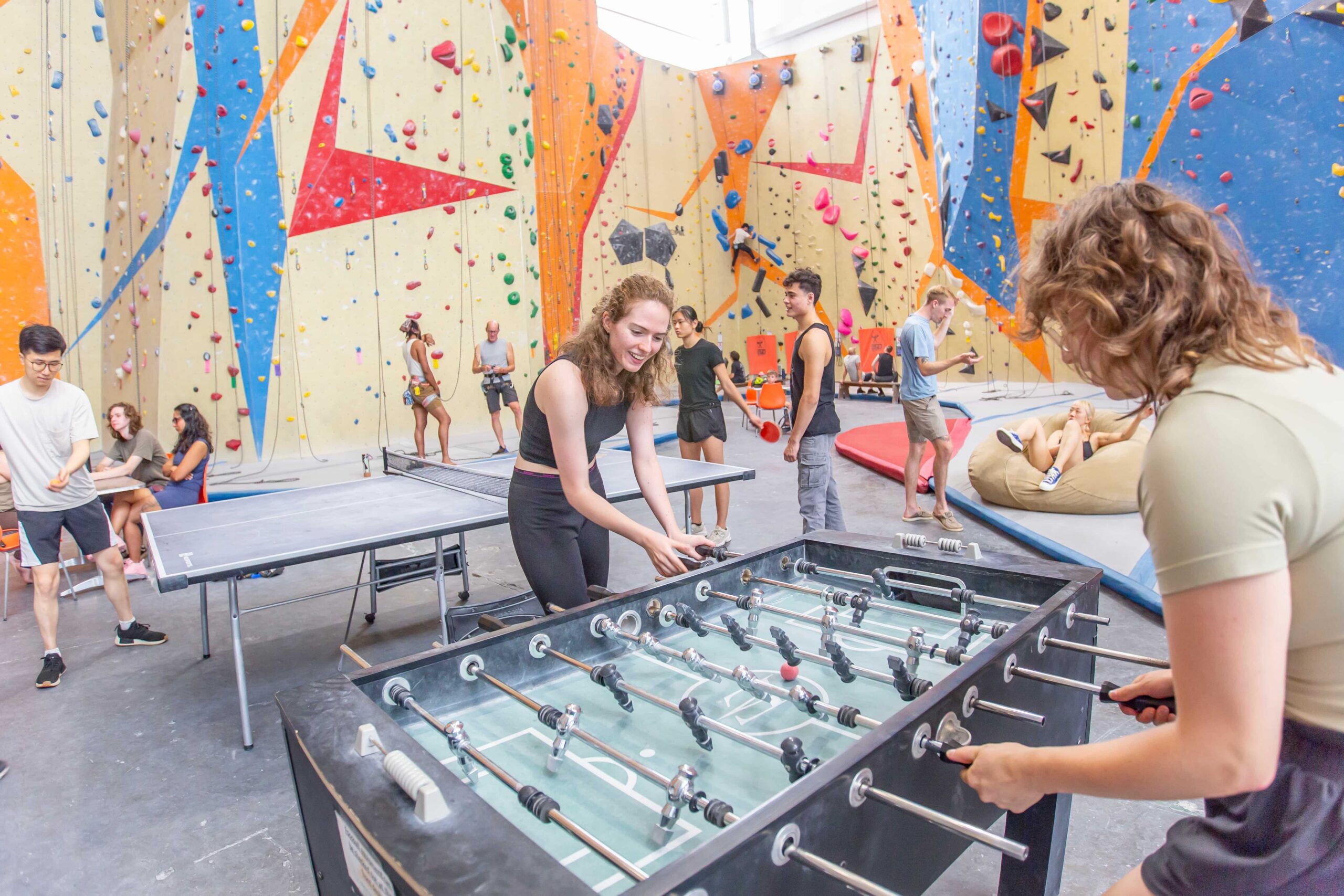 Two women play foosball together. Behind them people are playing ping pong and climbing.