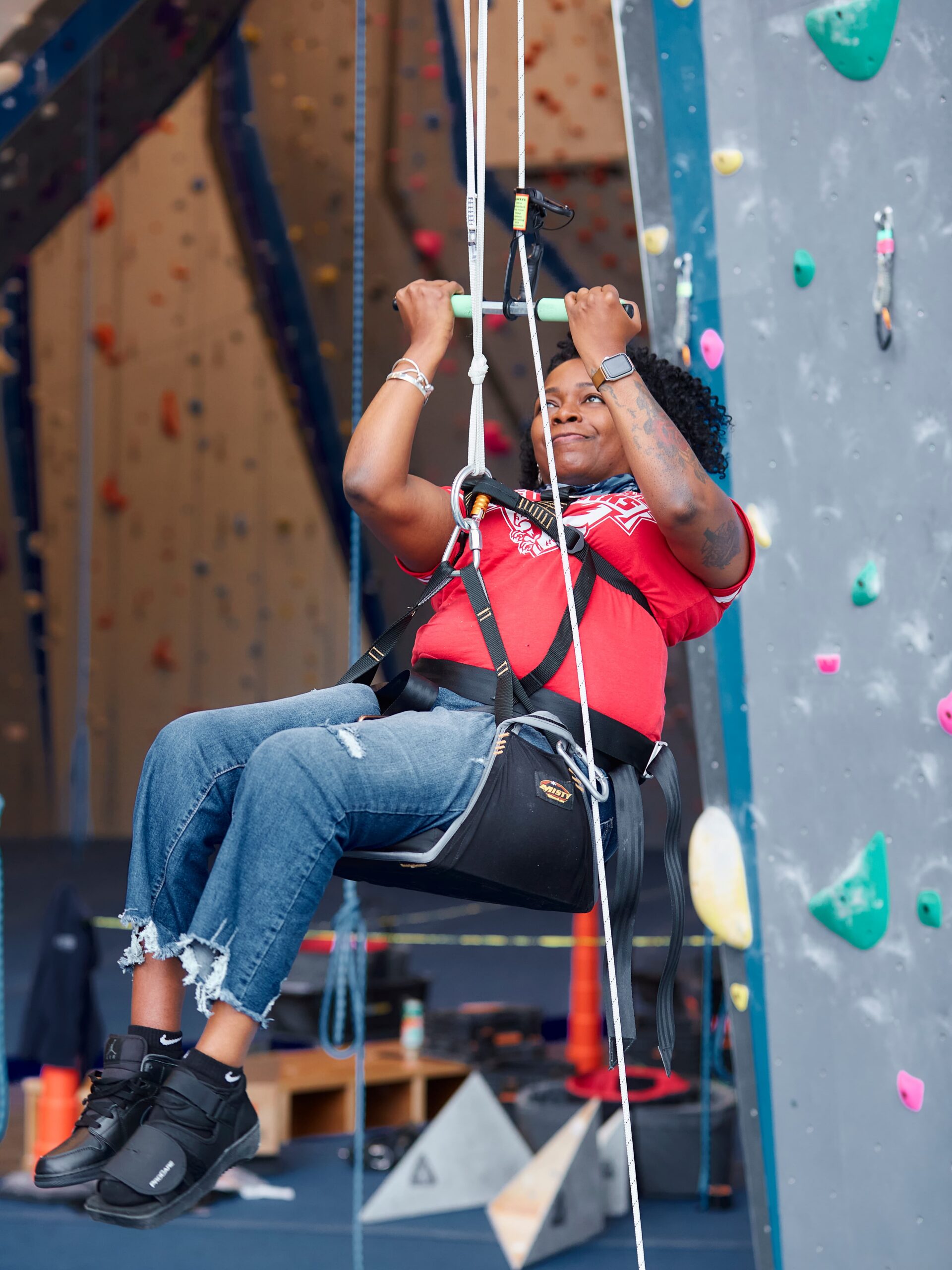 An adaptive climber ascends using a seat harness and pull up bar. She has tattoos and is wearing jeans and a t-shirt.