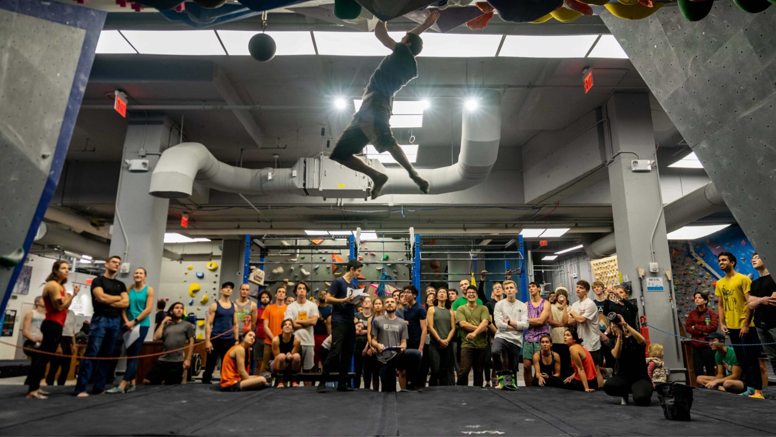 A climber campuses in the steep cave. Behind him, a crowd of people watch on.