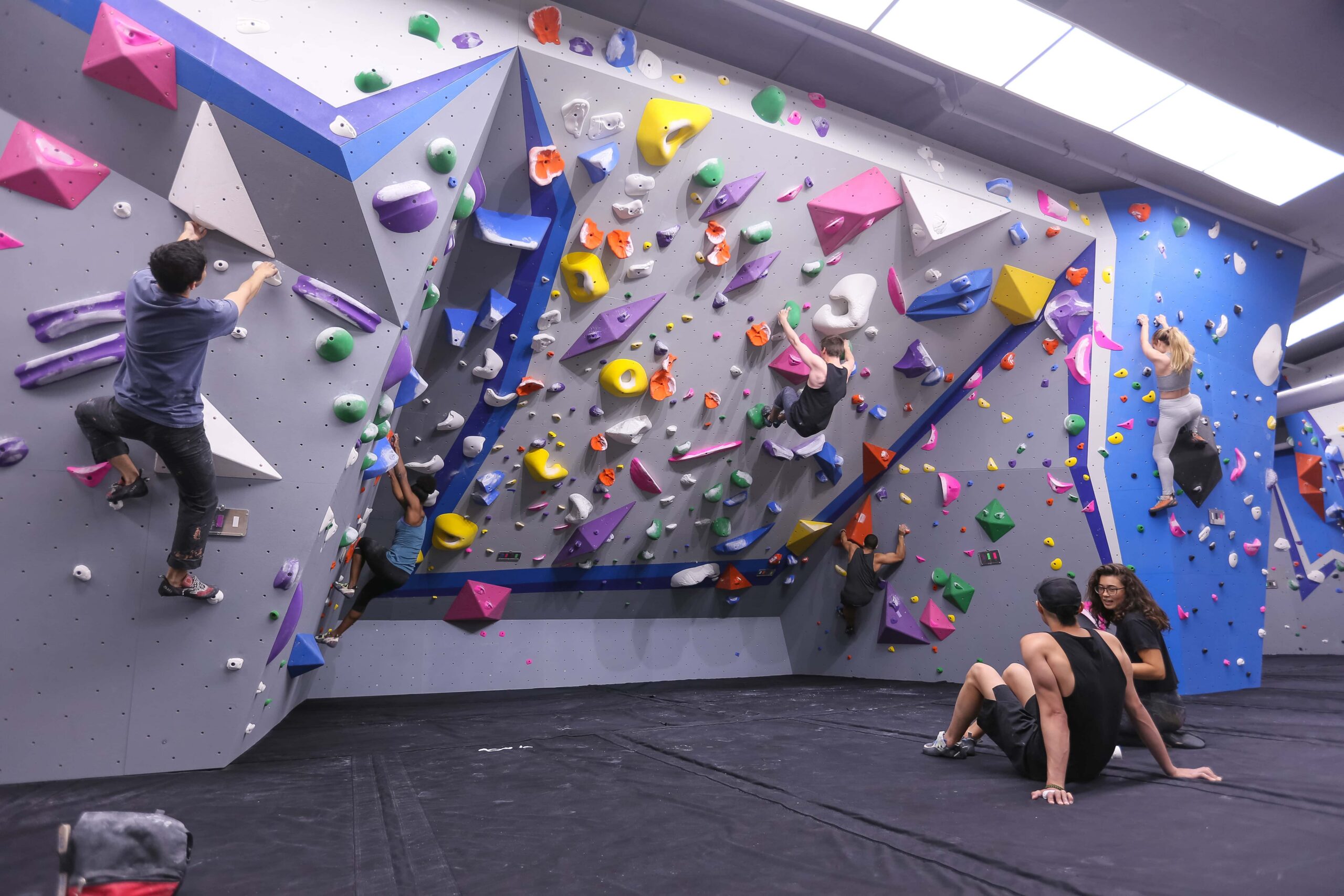 Two climbers sit on the mat and watch others boulder.