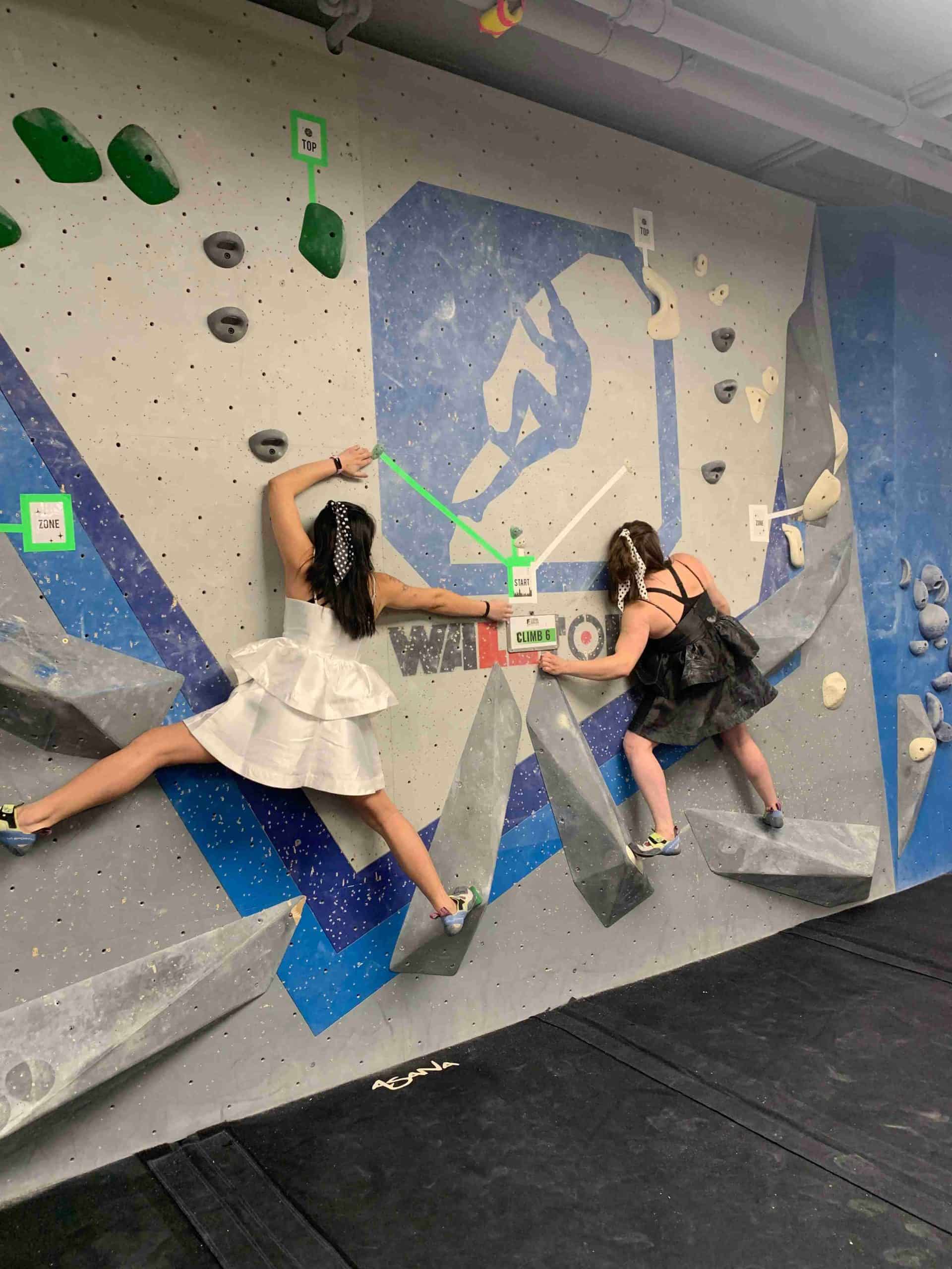Two girls wearing dresses are climbing a partner climb together. They are balancing on a slabby wall using volumes as feet.
