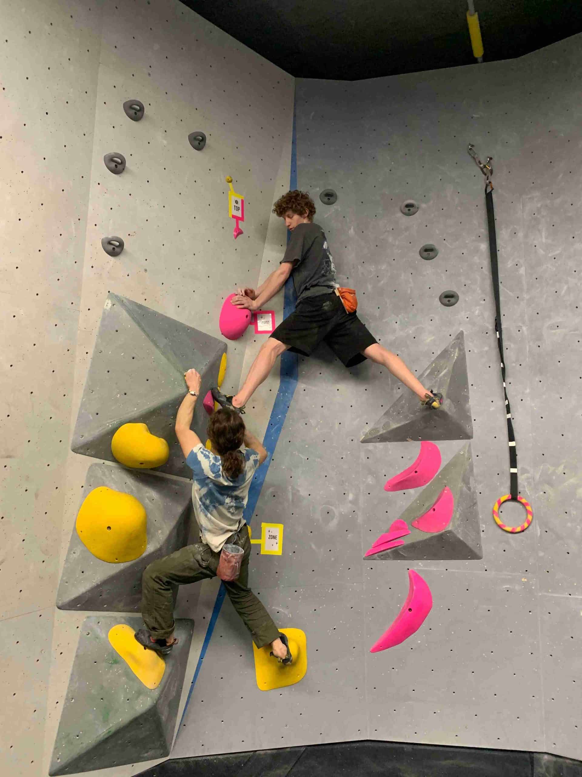 Two men complete a partner boulder together. On the climbers stands in a dihedral with each foot on a volume on the opposing walls. The second climber is grabbing hold of the first climber's foot as a hold.