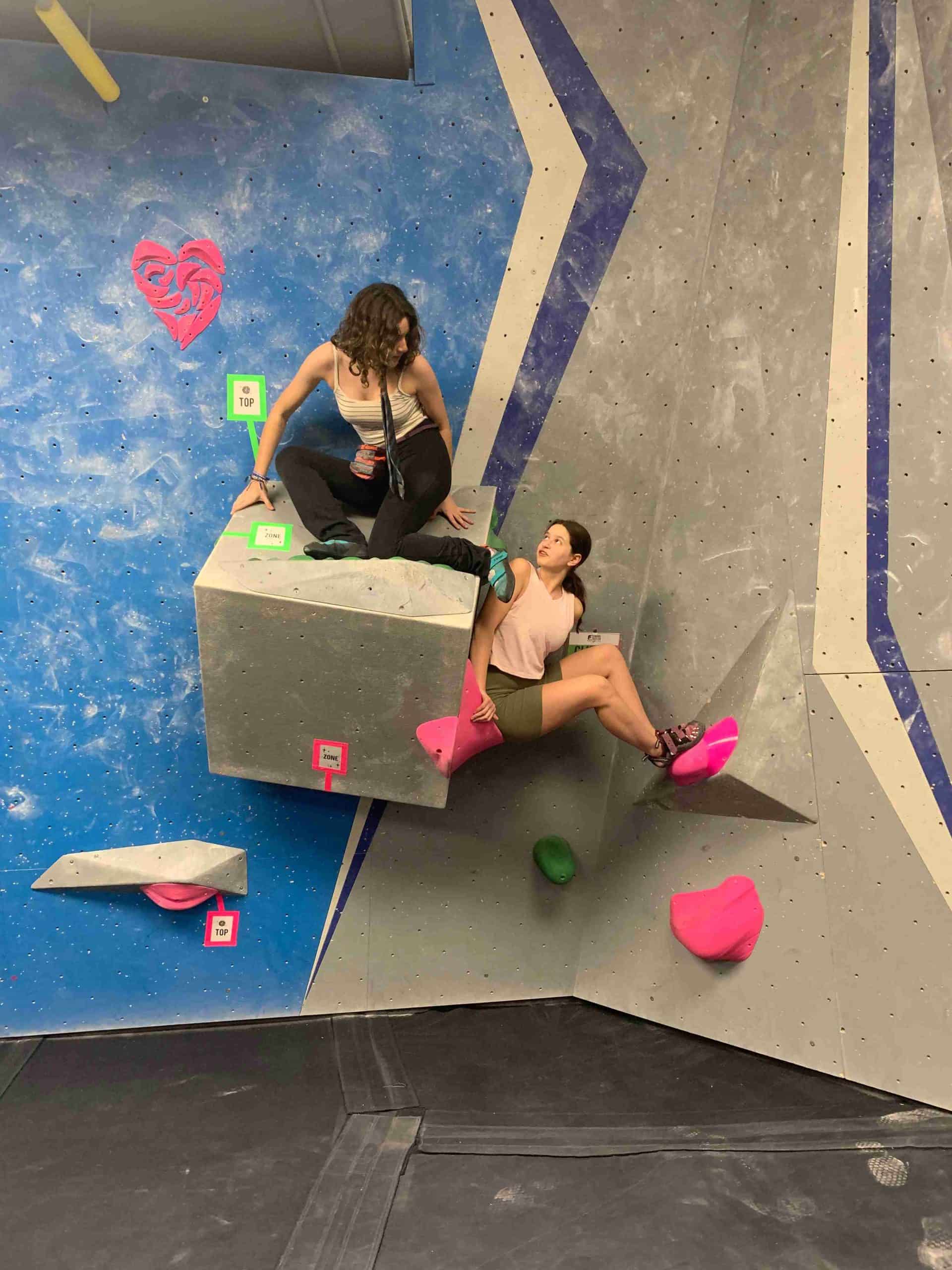 Two women climb a partner climb together. The first climber is wearing a tie and formal pants and is crouched on top of a square shaped volume with her back to the wall. The second climber is resting her back against the square volume, resting her feet on another volume, and looking back and up at the first climber.