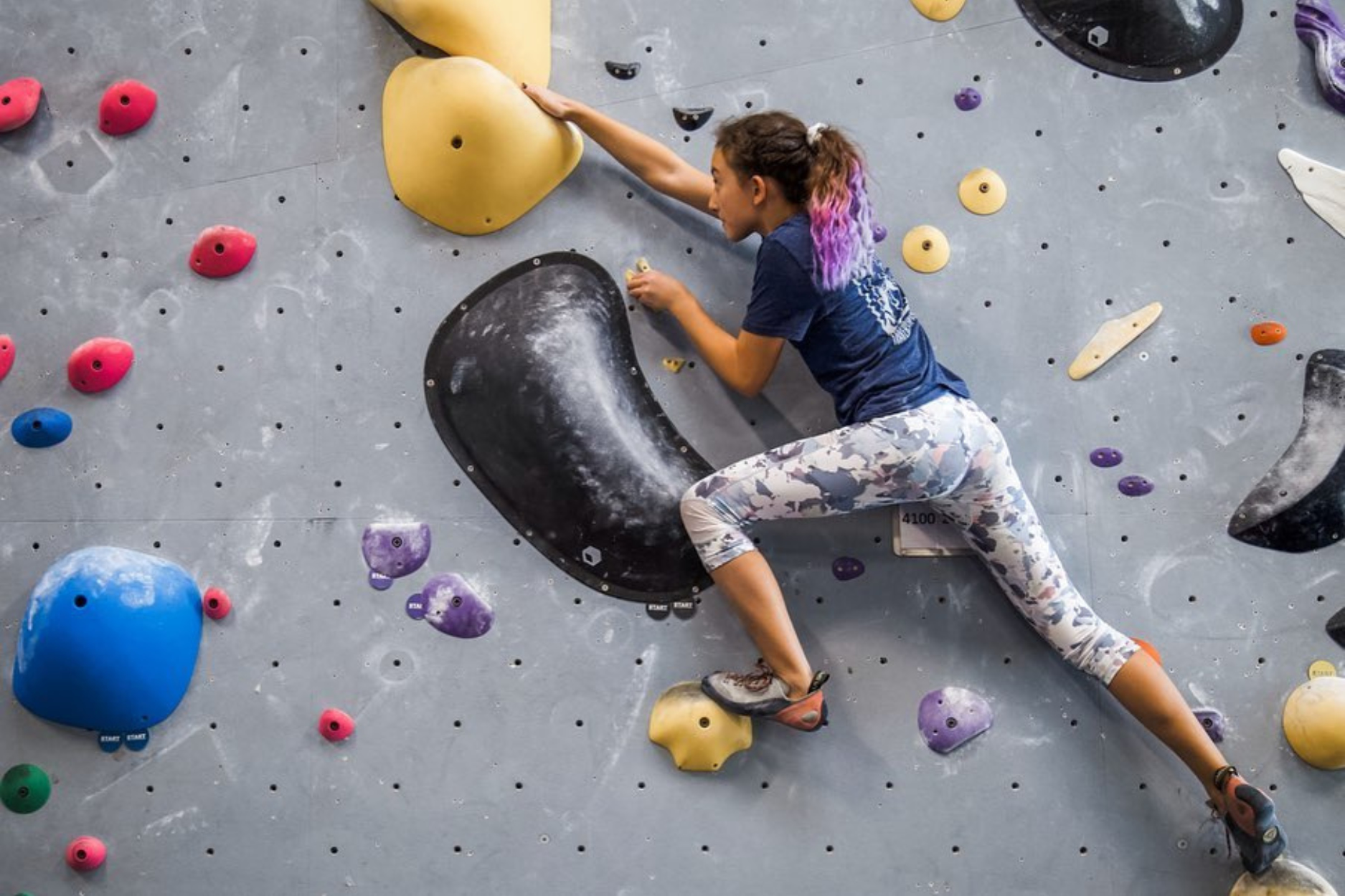 A young girl is bouldering and reaching for the next hold on the wall.