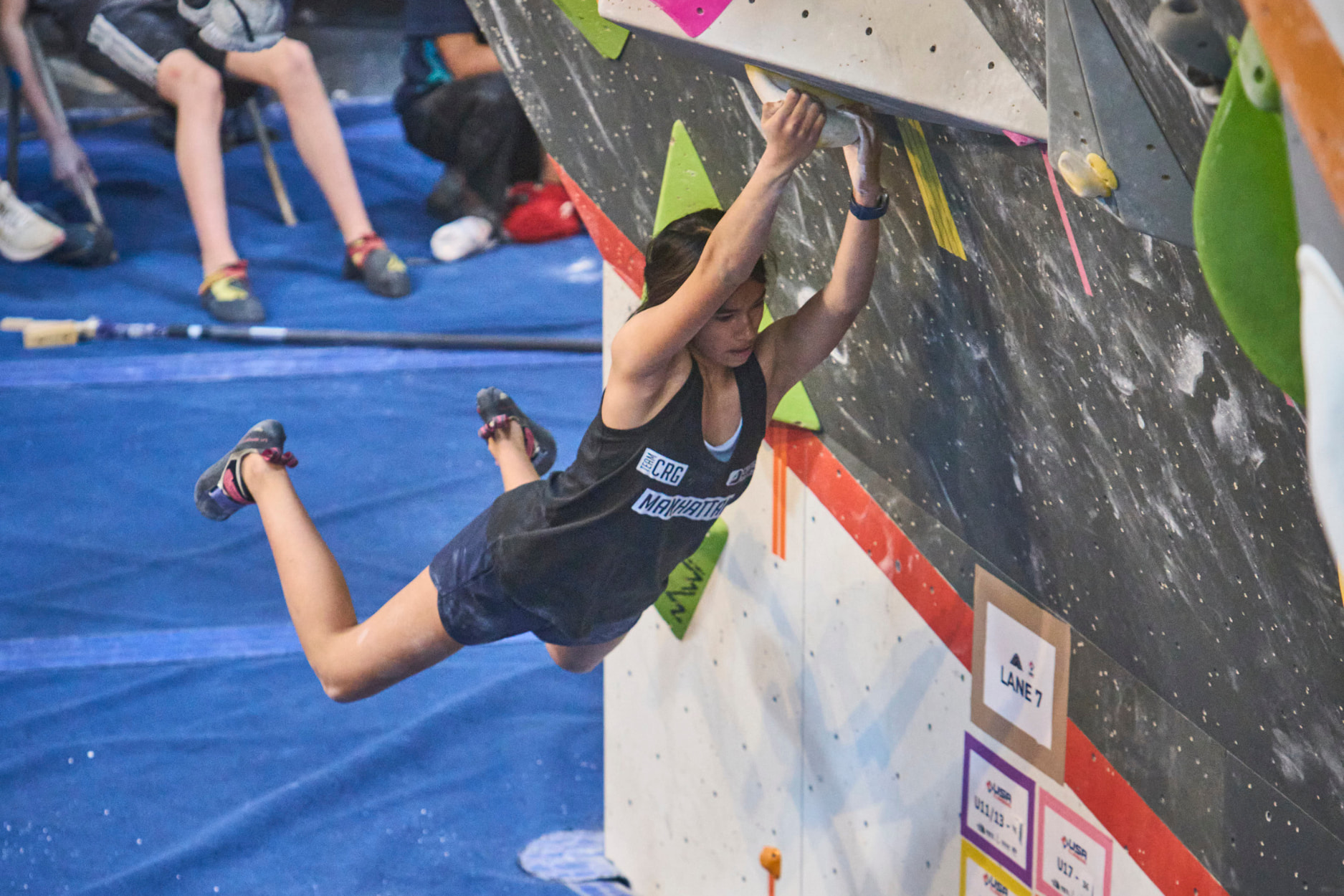 A girl wearing a Team CRG Jersey kicks her feet back to prepare for a lache to the next hold in a bouldering competition.