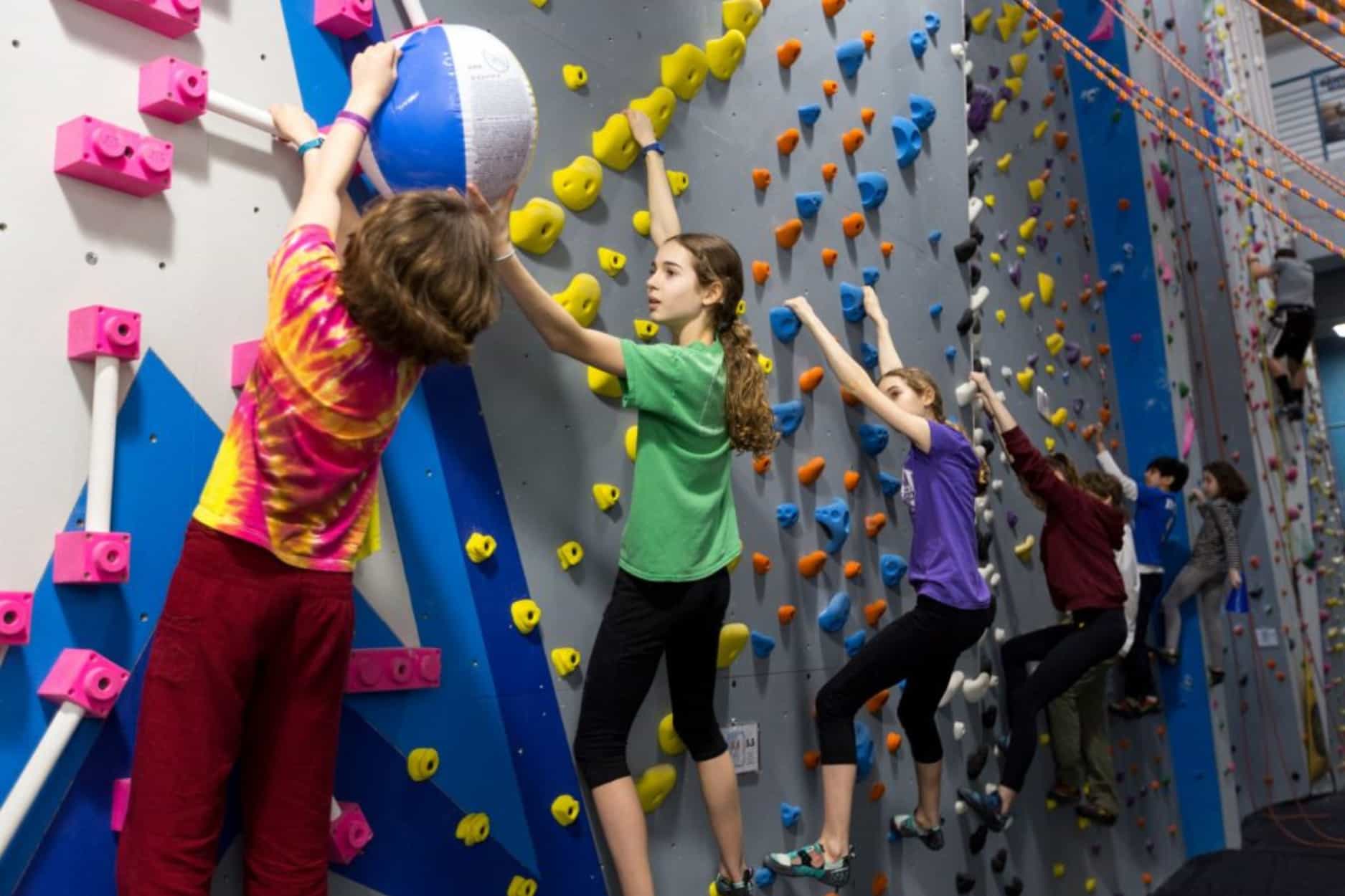 A group of kids playing a traversing challenge game at a youth program where they hang onto the climbing wall and pass a beach ball down the line without falling off the wall.