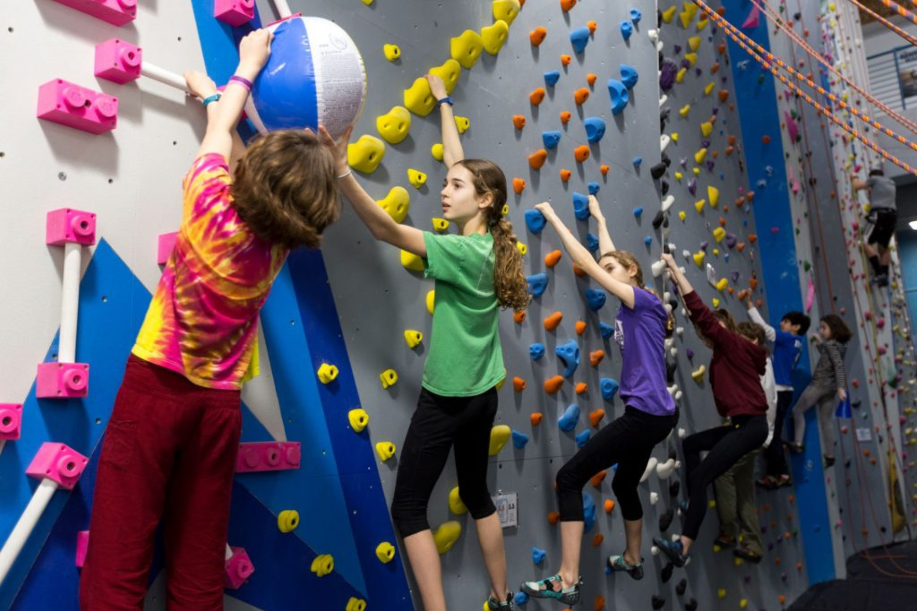 A group of kids playing a traversing challenge game at a youth program where they hang onto the climbing wall and pass a beach ball down the line without falling off the wall.